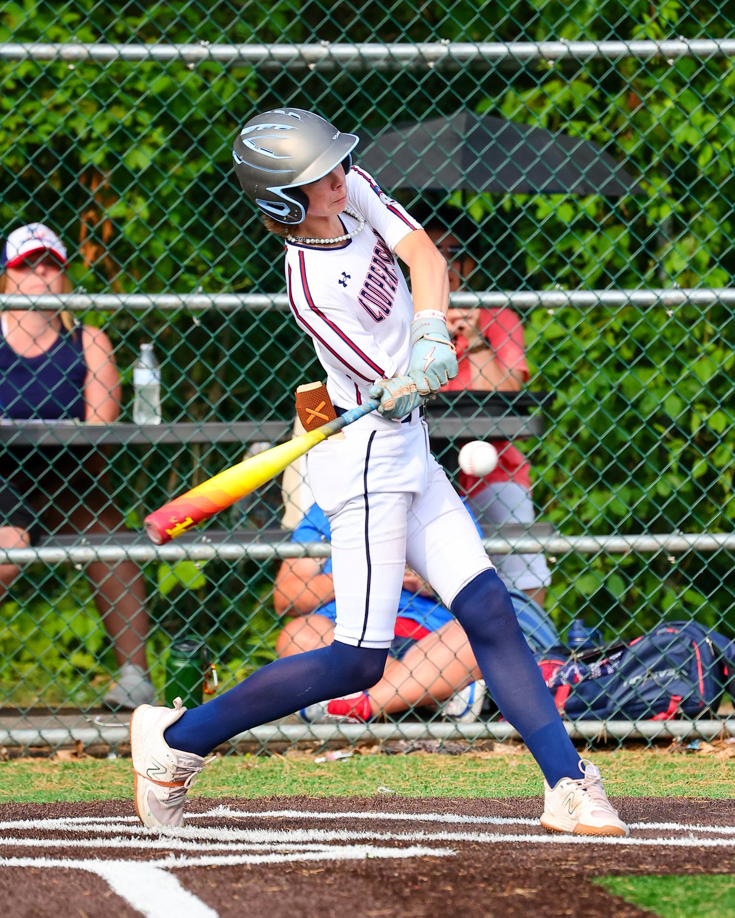 A baseball player in navy and white uniform swings the bat at home plate with green foliage in the background.