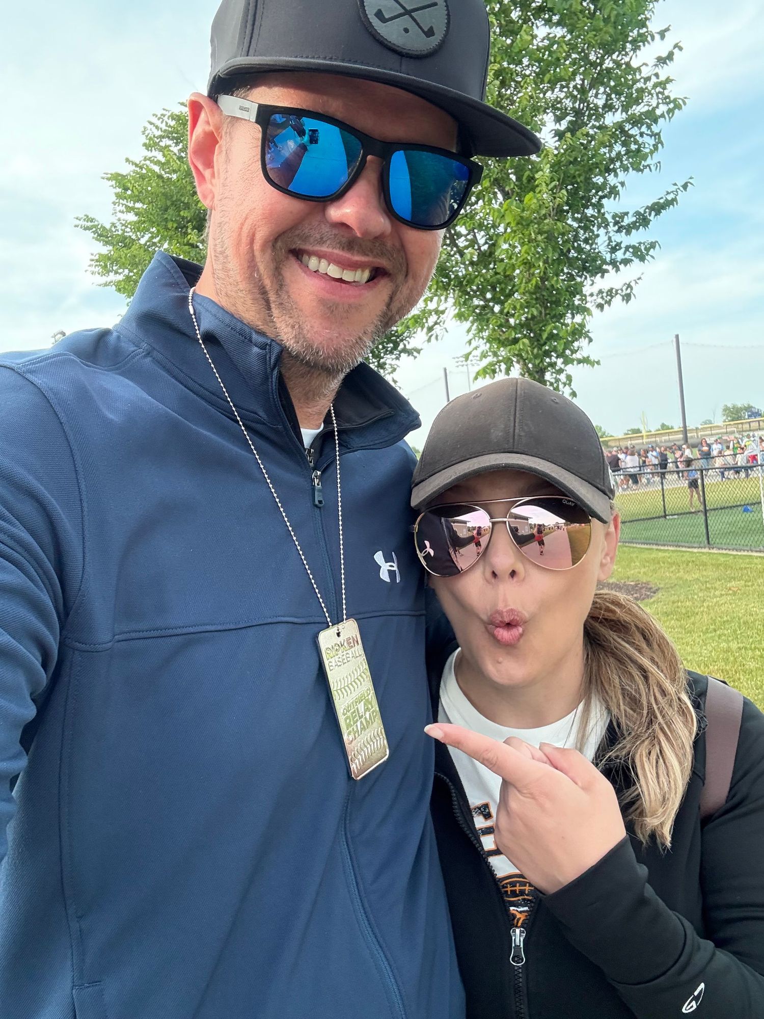 Two spectators wearing baseball hats and sunglasses posing together at a sports event on a sunny day.