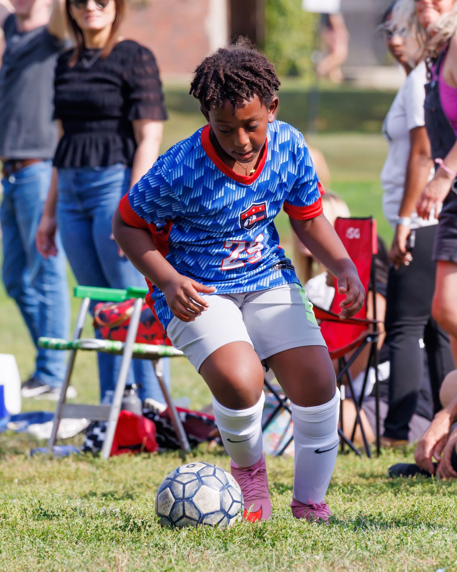 Young soccer player in blue jersey #23 dribbles ball on grass field during outdoor game with spectators watching.