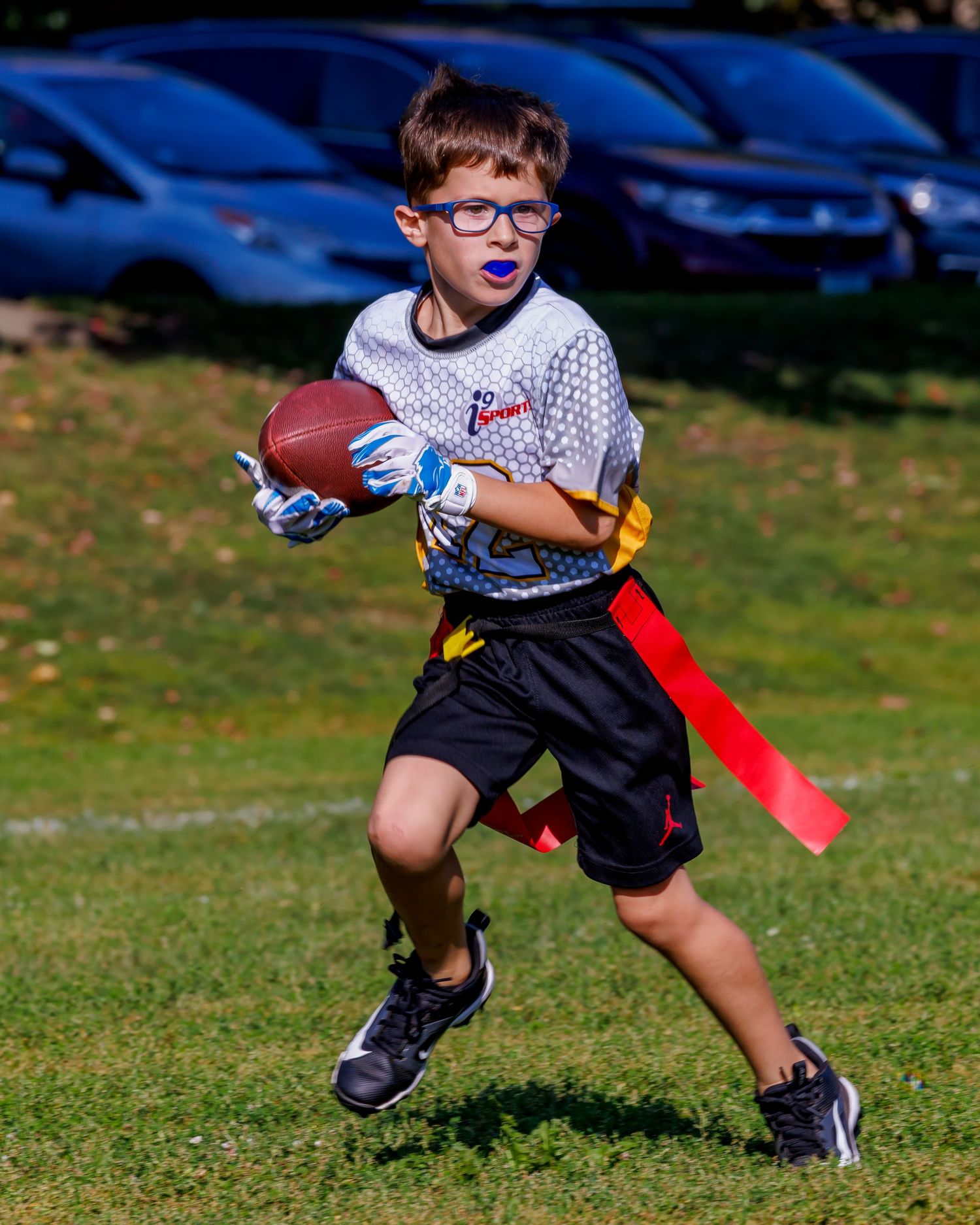 Young boy in glasses playing flag football, running with ball wearing blue gloves and red flags on sunny field.