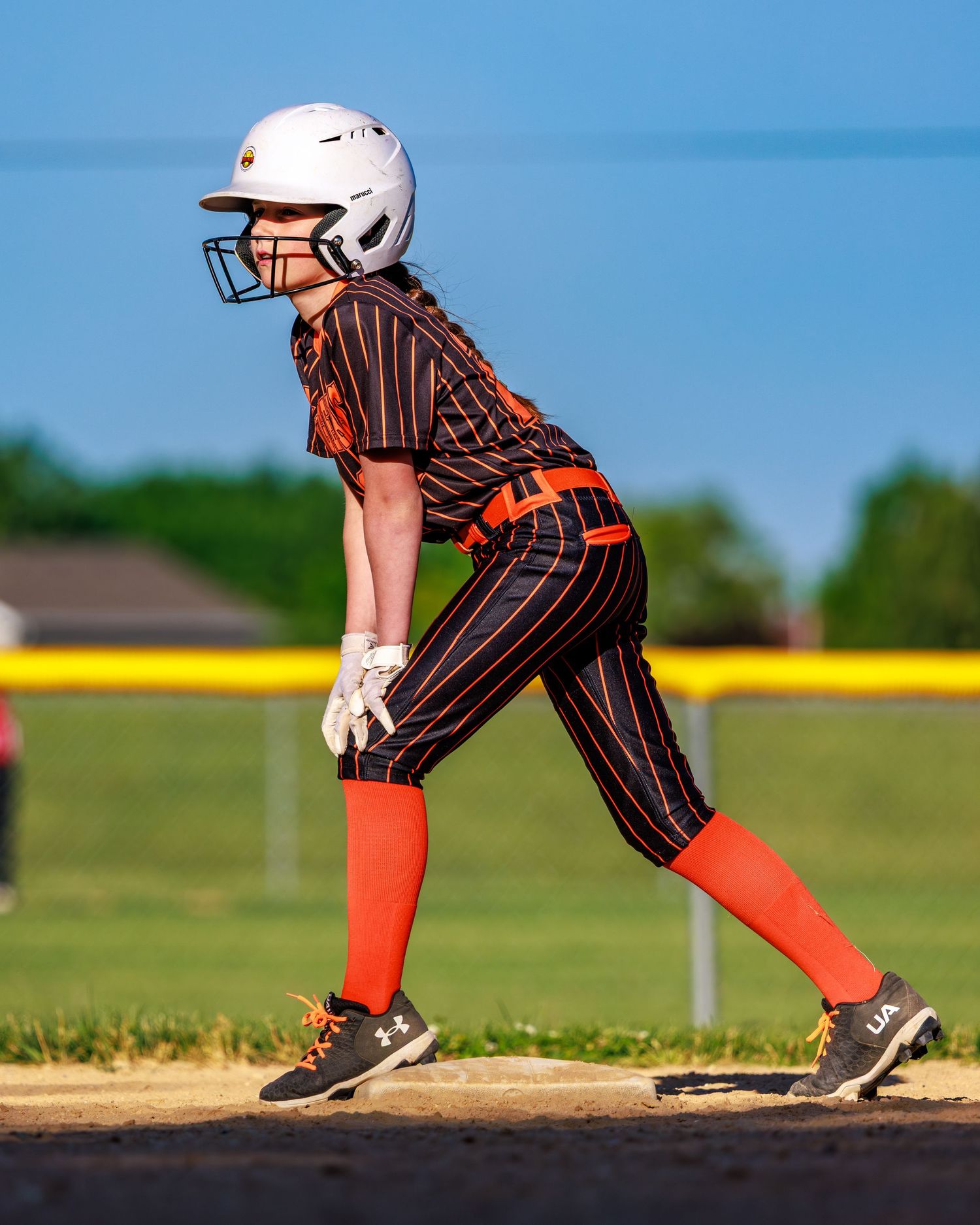 Young softball player in orange and black pinstripe uniform ready at base during a game.
