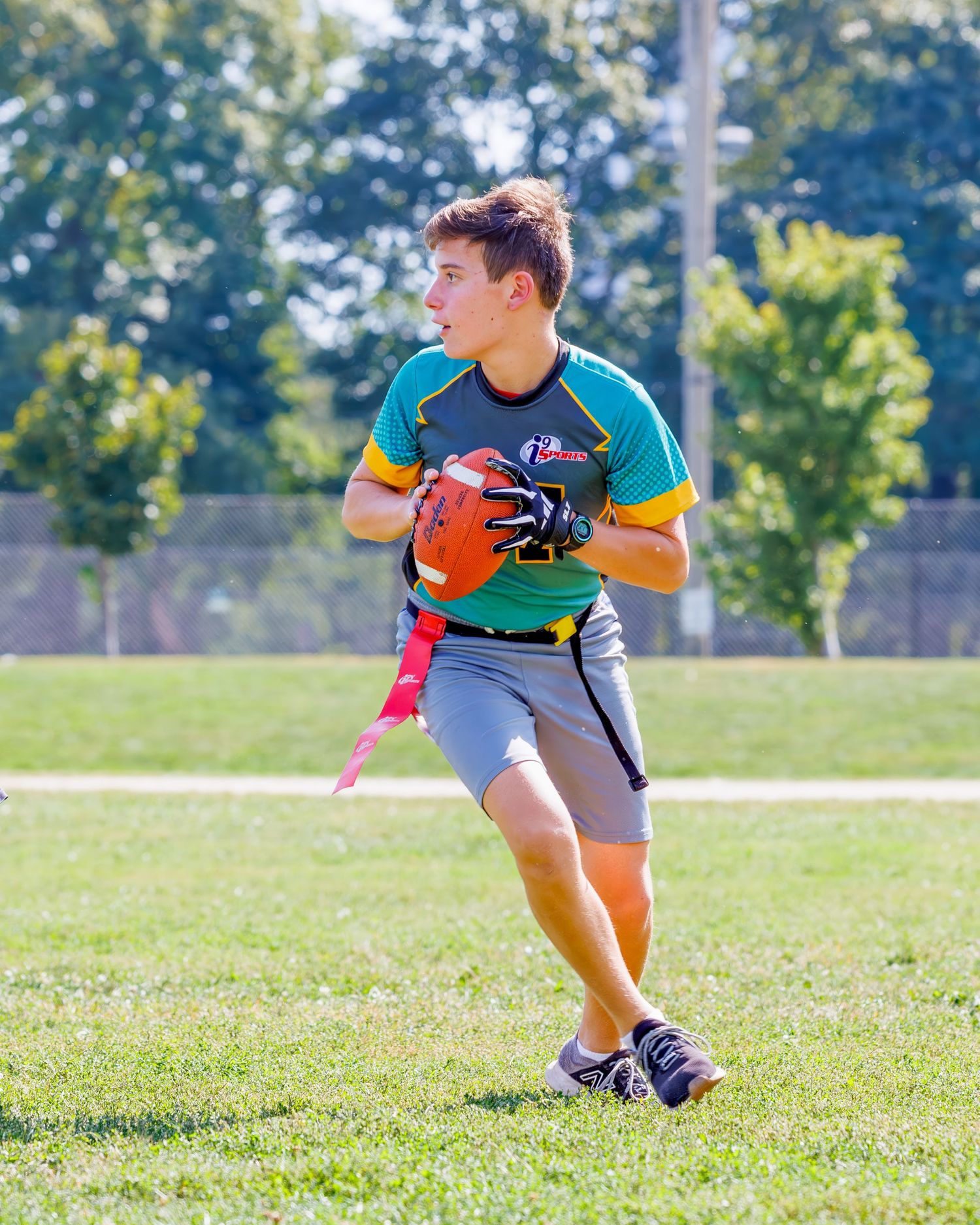 Flag football player in teal and yellow jersey runs with ball on sunny grass field during game action.
