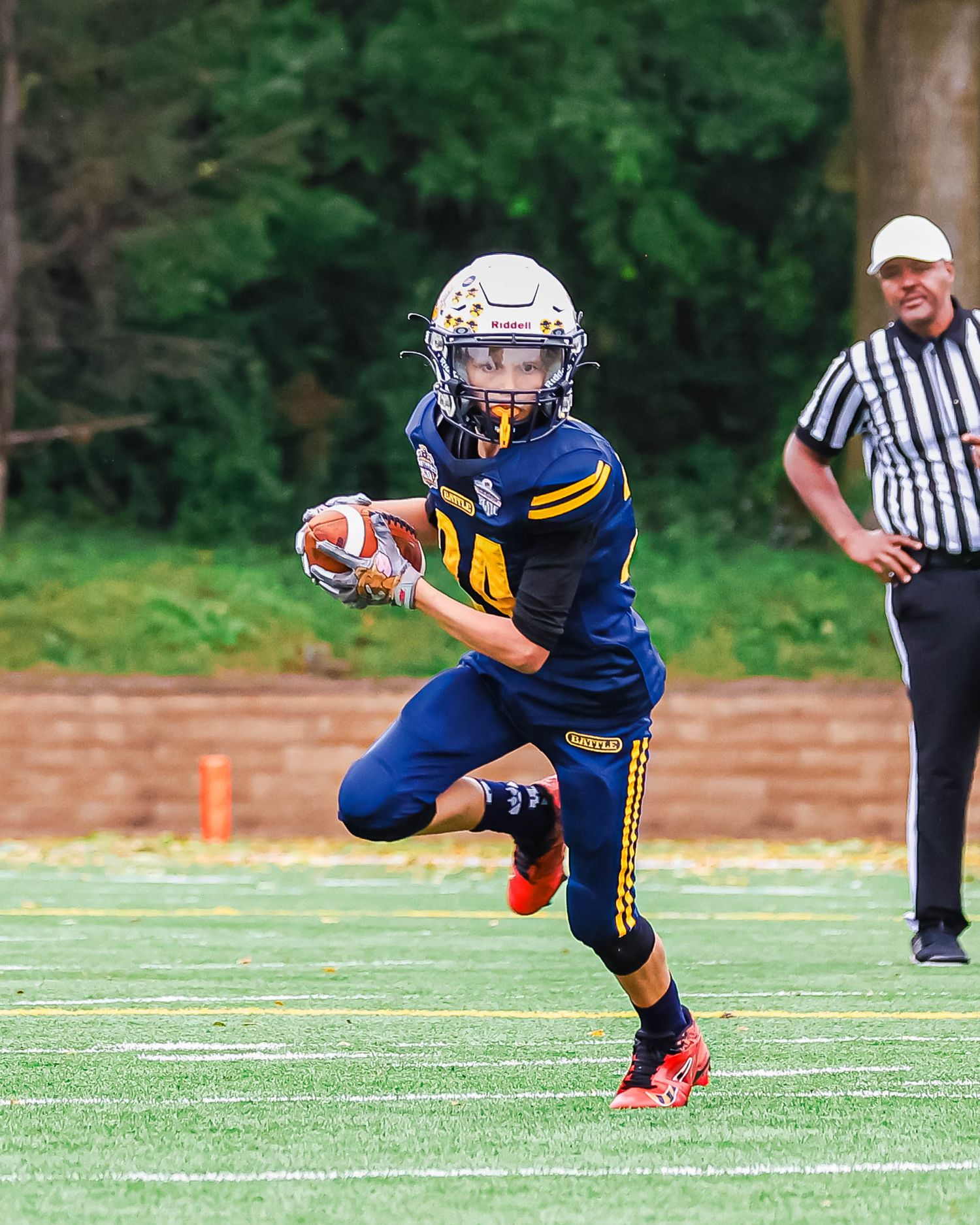Football player in blue jersey runs with ball on turf field while referee watches in background.