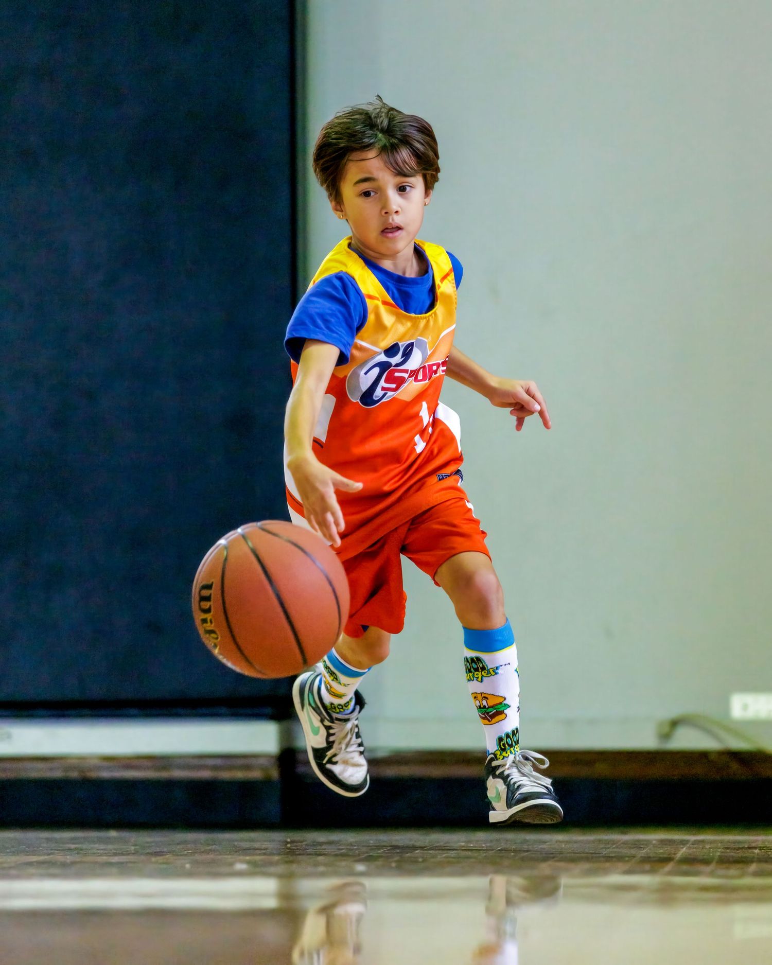 Young boy in orange and blue basketball uniform dribbling a Wilson basketball on an indoor court.
