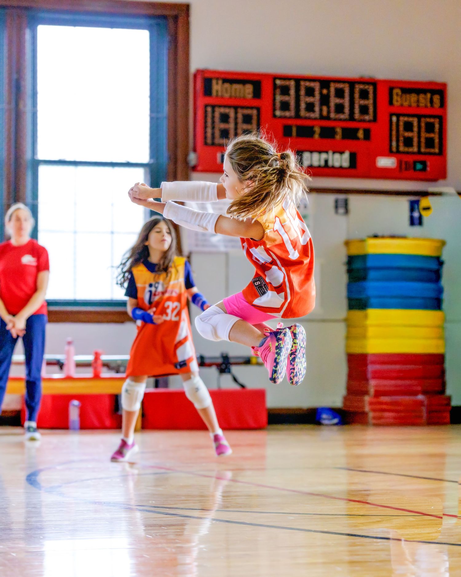 Young athletes practice high kicks in a colorful gymnasium with red scoreboards and safety mats on the floor.