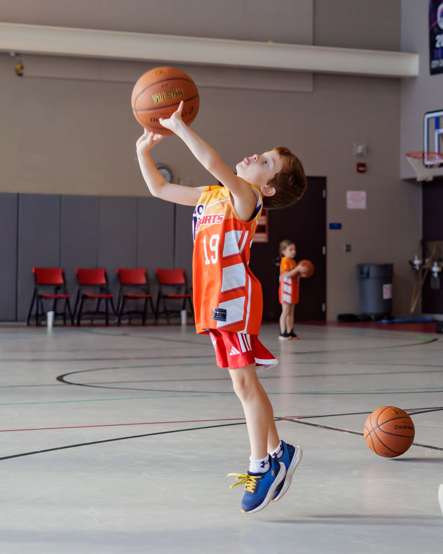 Basketball player in orange jersey practices dribbling and shooting drills in an indoor gymnasium.