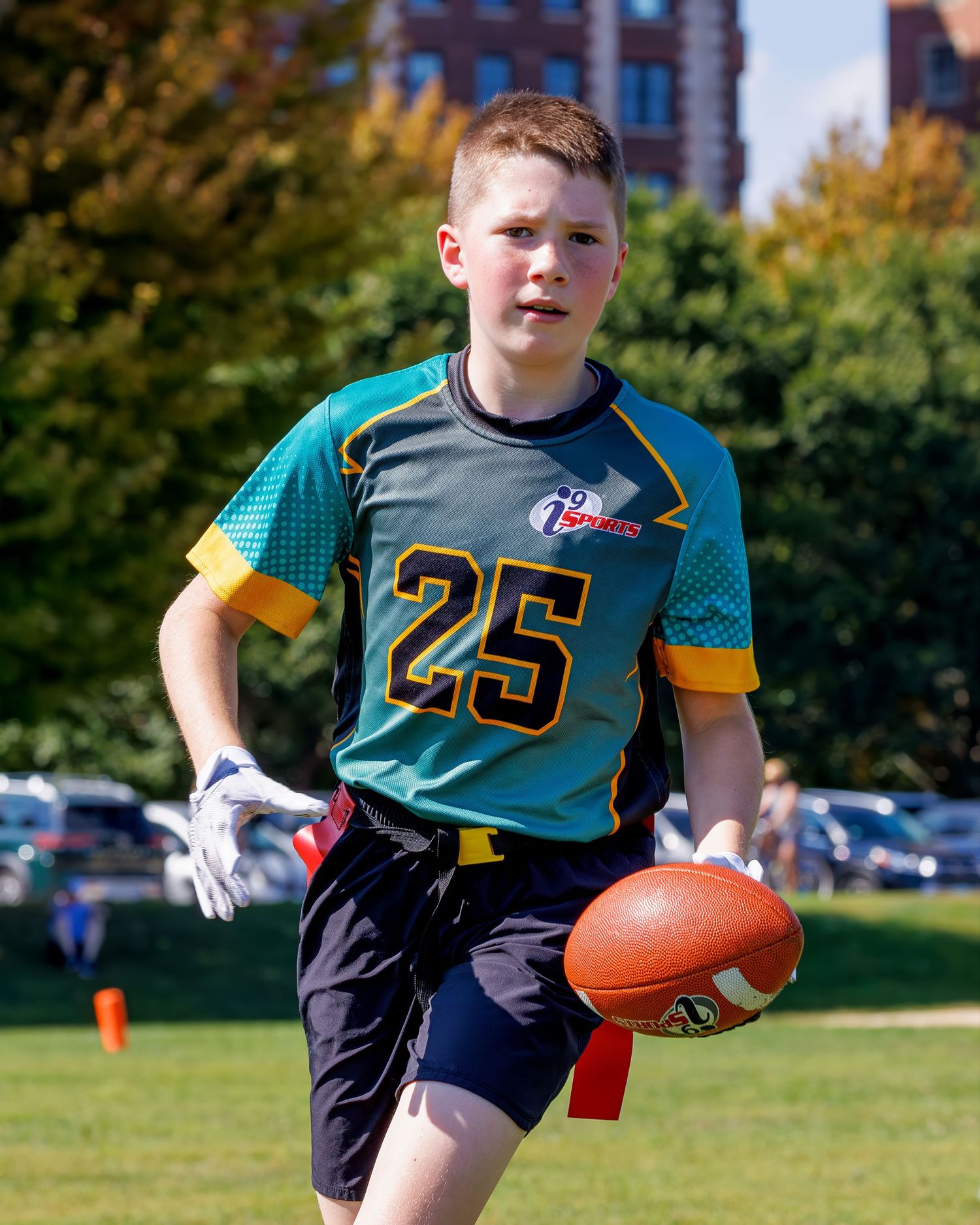 Youth flag football players in teal and yellow uniforms run with footballs during practice drills.