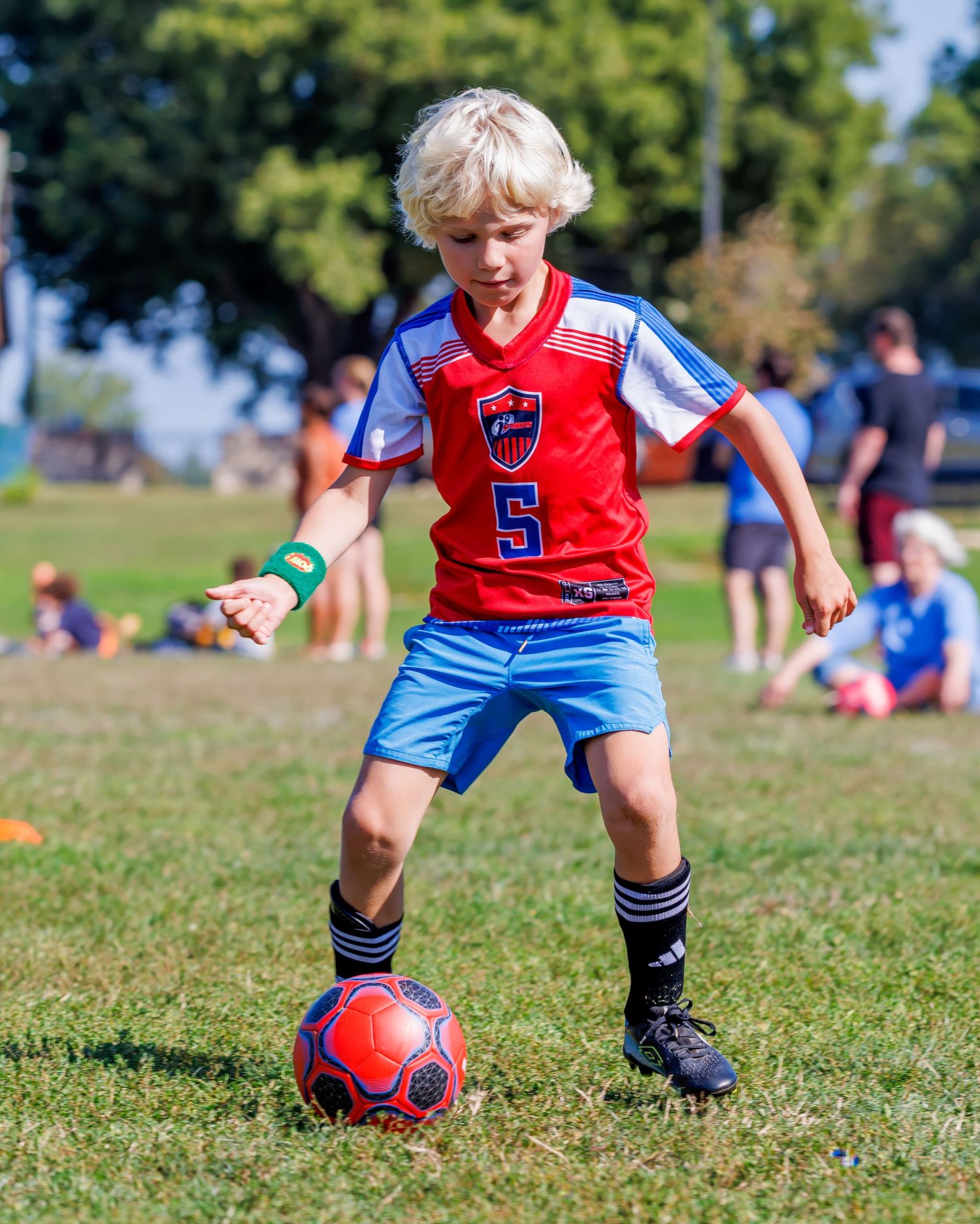 Young blond boy in red number 5 soccer jersey dribbling a red ball on green grass field during youth soccer game.