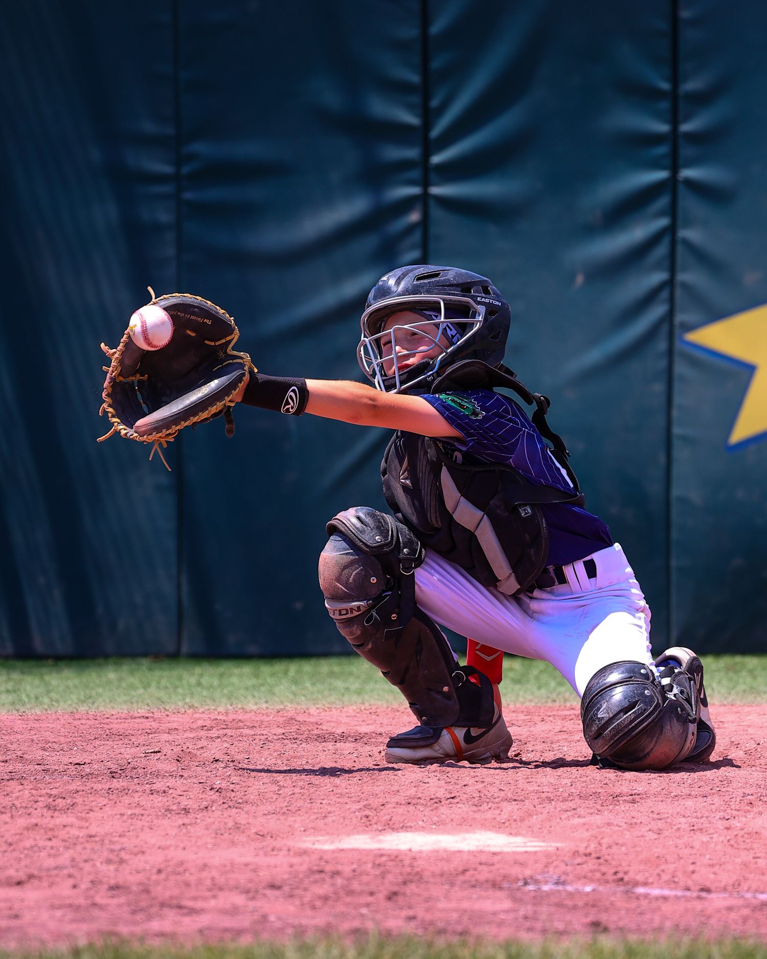 Baseball catcher in protective gear crouches in ready position behind home plate during game.