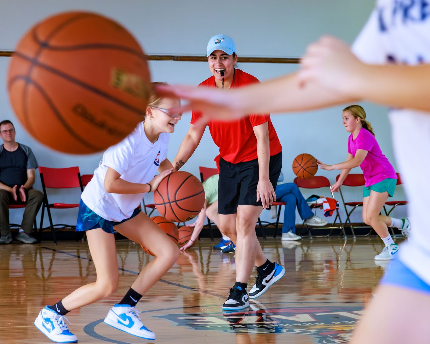 Young athletes practice basketball dribbling drills in a gymnasium with a coach in a red shirt supervising.