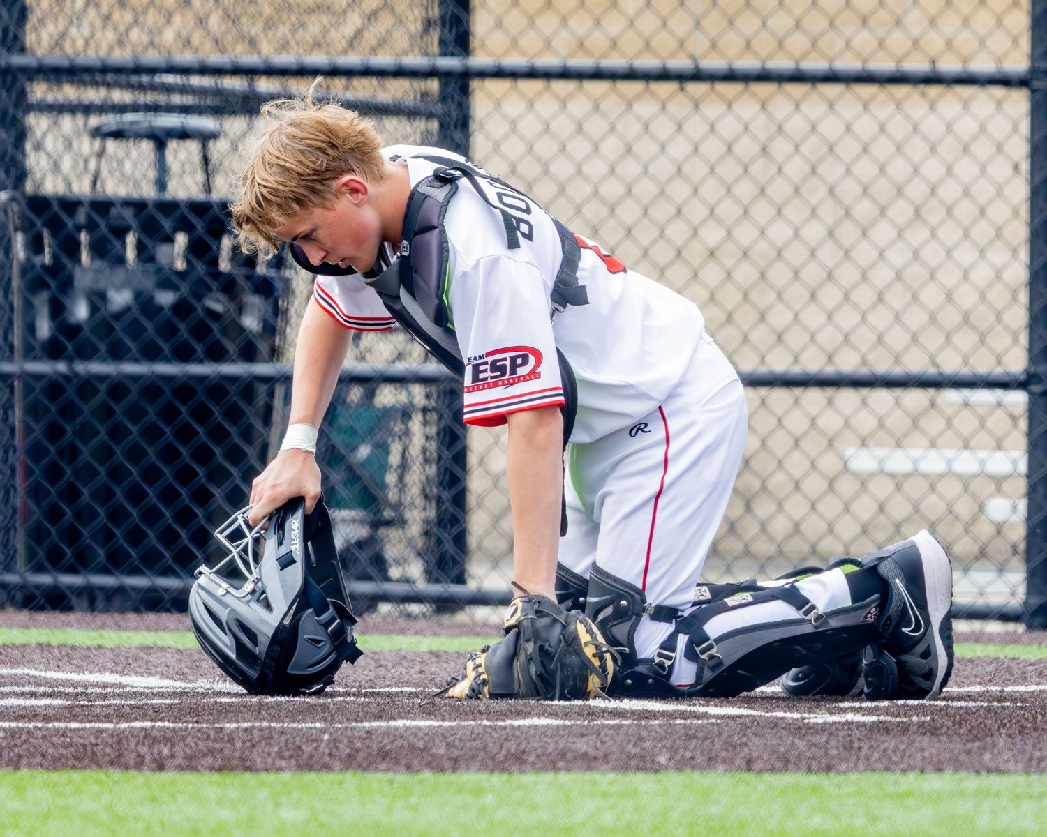 Baseball catcher in white uniform kneeling to adjust equipment on home plate during game.