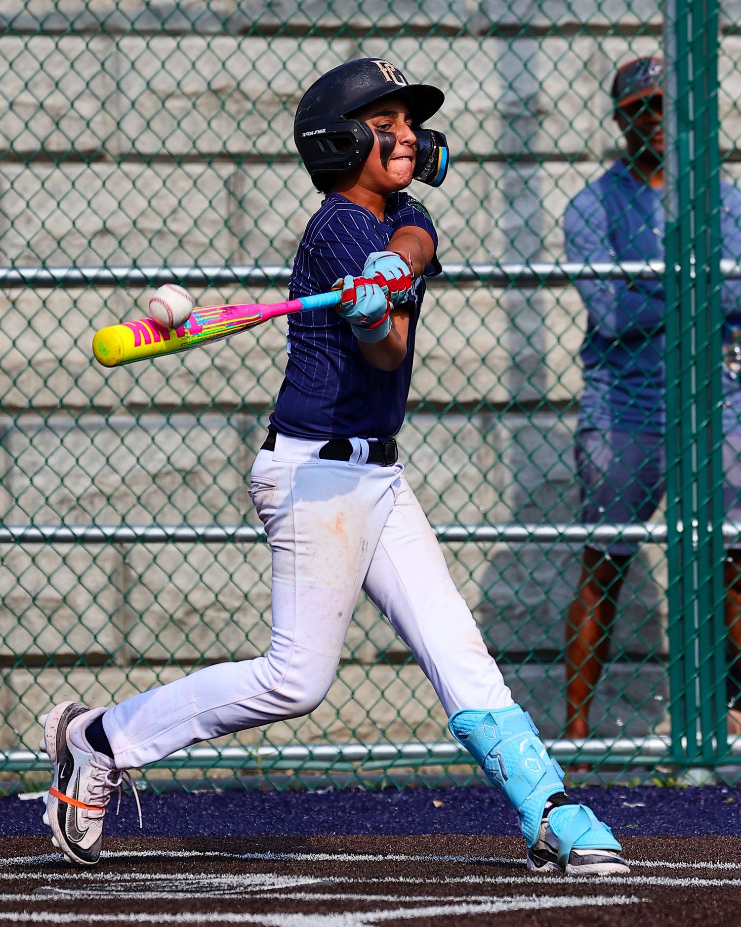 Youth baseball player in navy uniform and teal socks swings a colorful bat during a game at a fenced field.