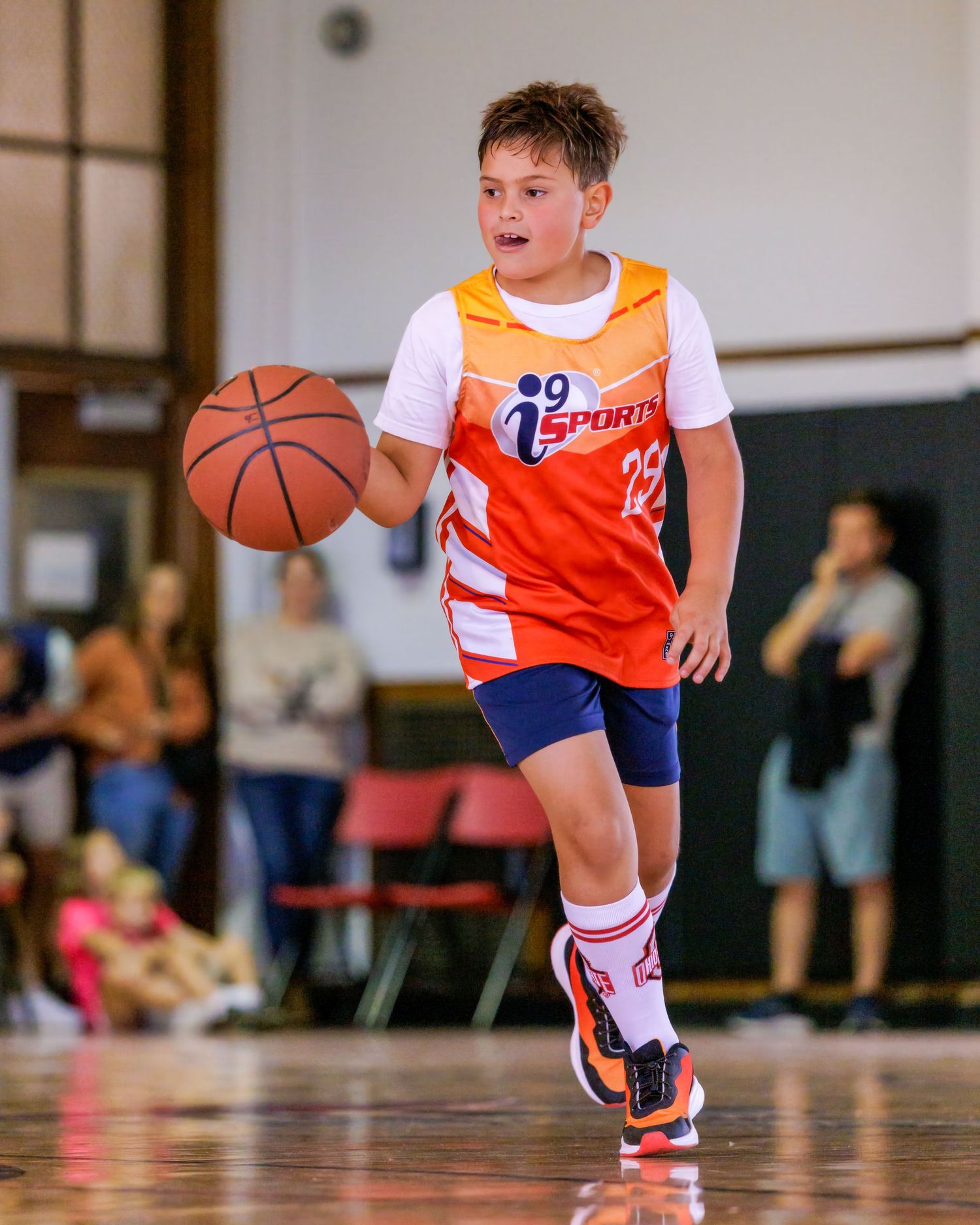 Young boy in orange iSports jersey dribbling basketball on indoor court during youth league game.
