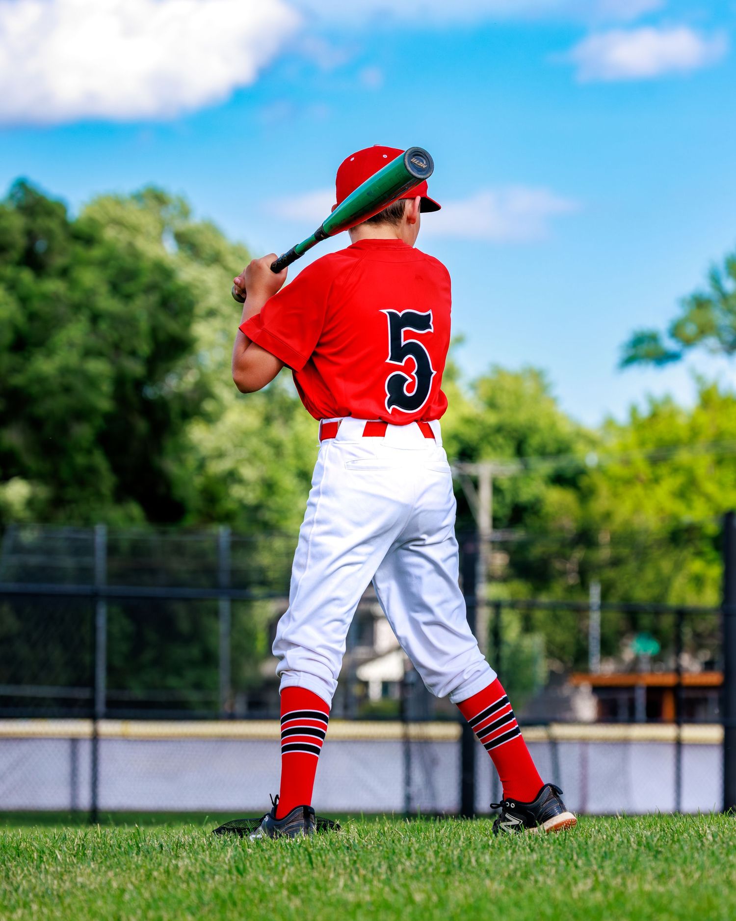 Young baseball player in red uniform number 5 stands ready to bat on a sunny green field.