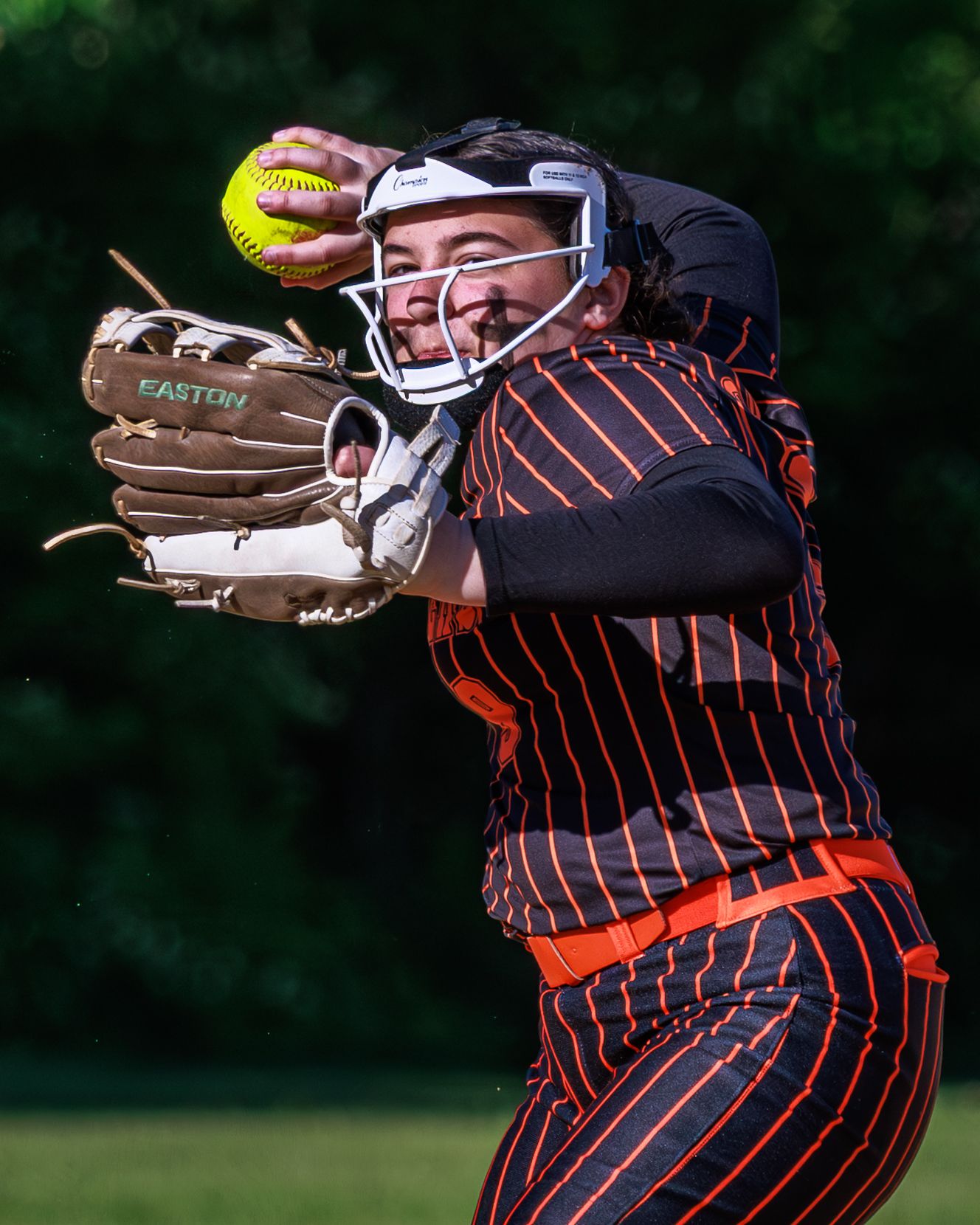 Softball player in black and orange striped uniform with Easton glove catching a yellow softball while wearing a catcher's mask.