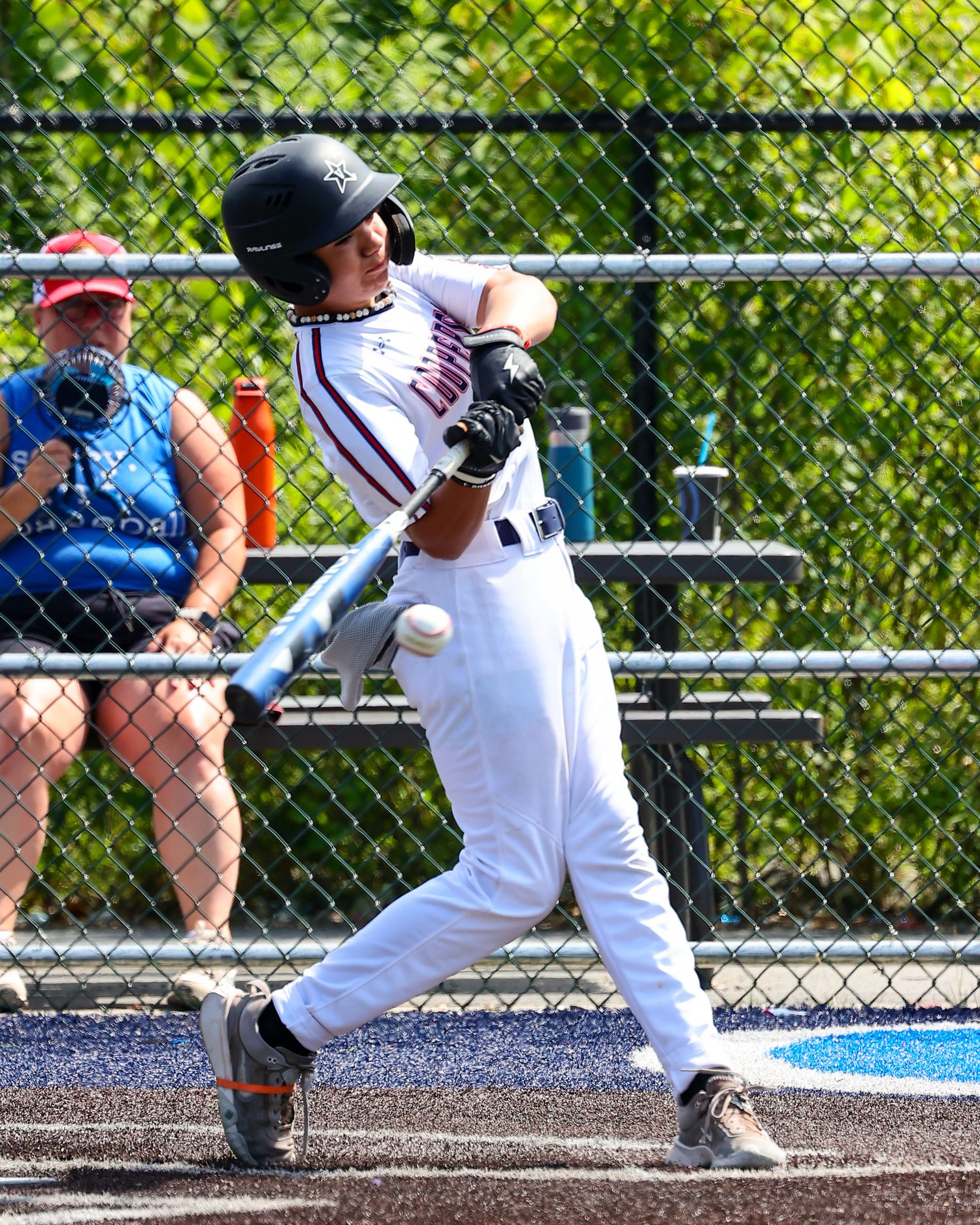 Baseball player in white uniform swings bat in sequence showing follow-through motion against chain link fence background.