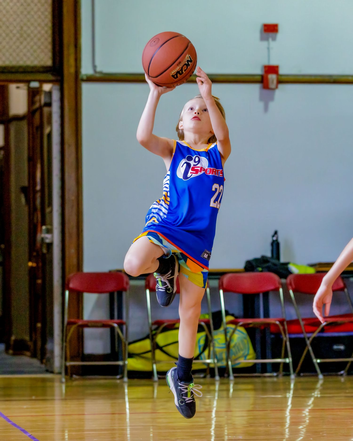 Young basketball player in blue jersey jumps high while shooting the ball during a game in a gymnasium.