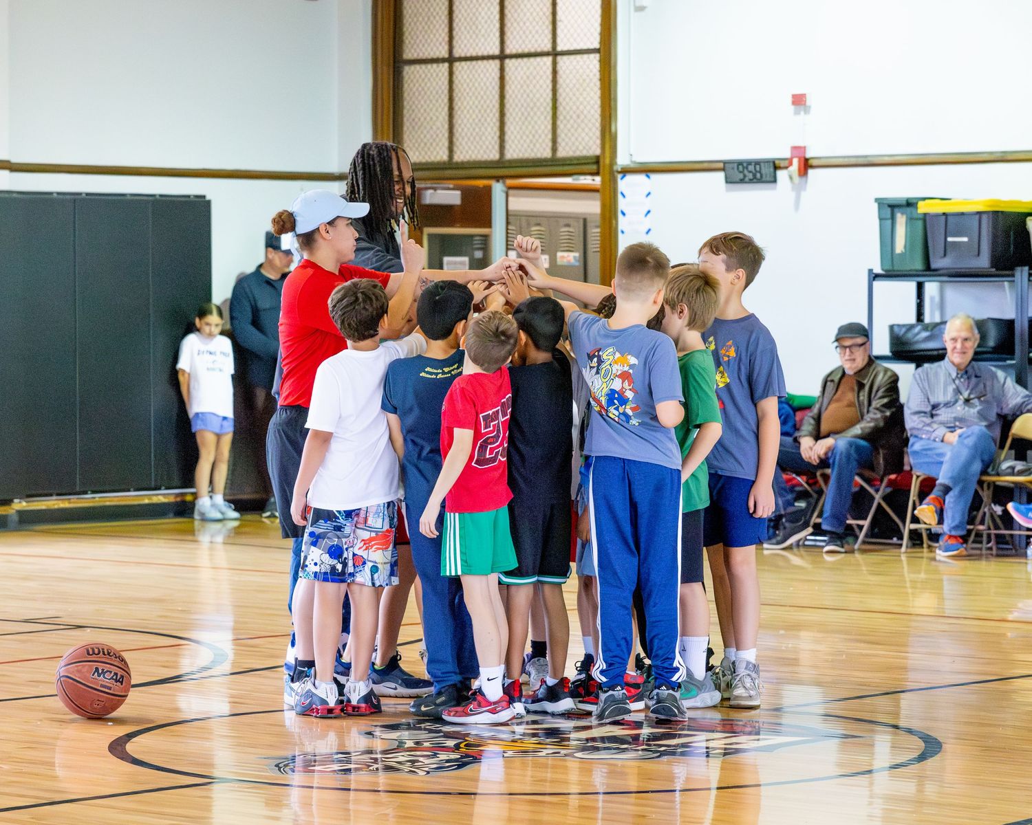 Students gather in a gymnasium for basketball practice while receiving instruction from coaches on the court.