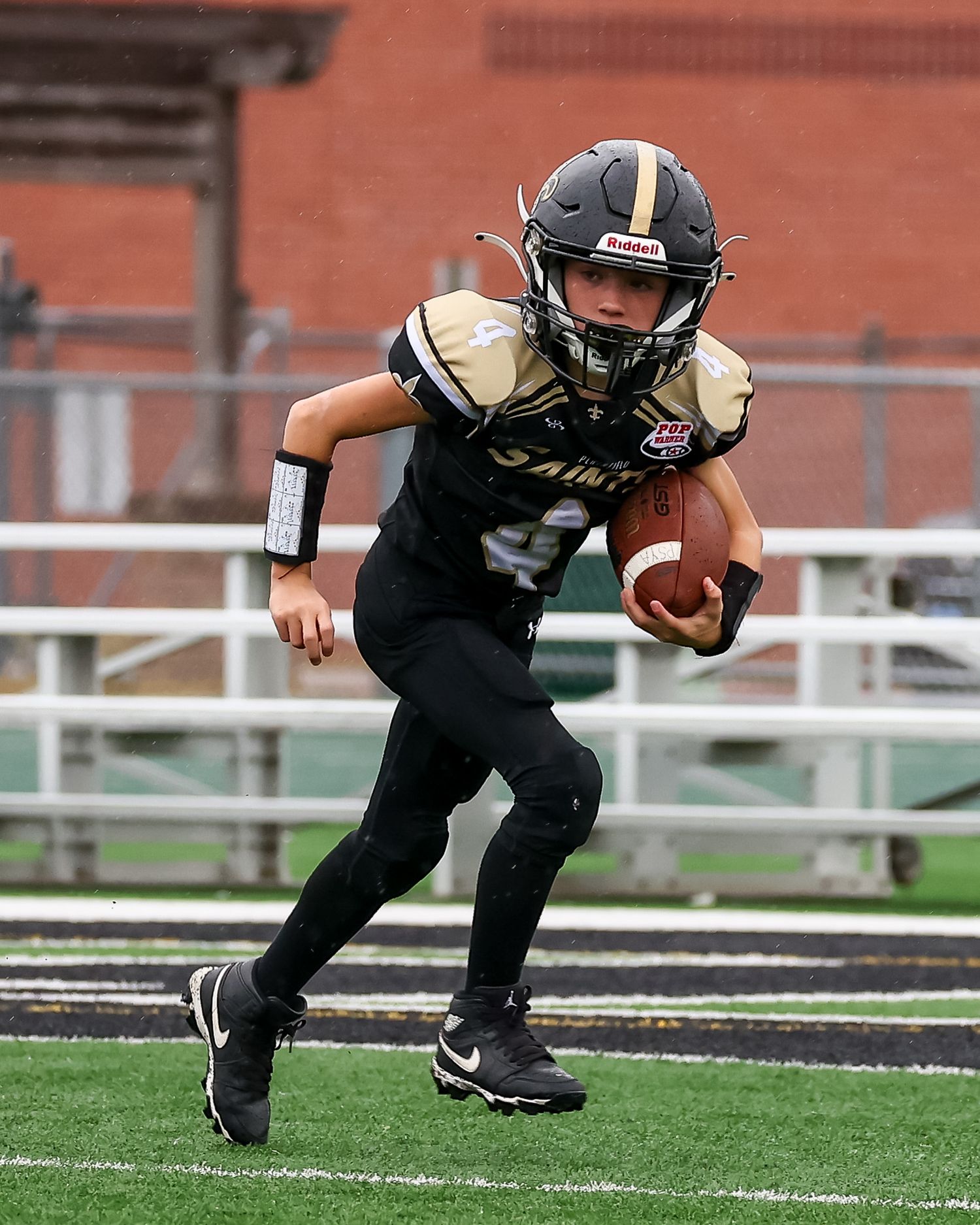 Young football player in black Saints uniform runs with ball during youth league game on turf field.