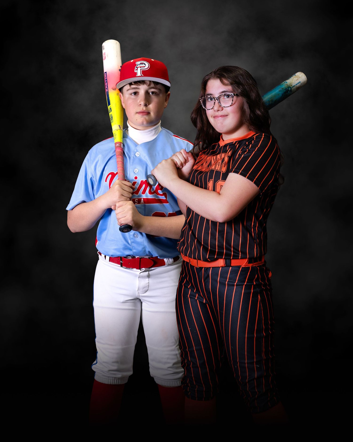 Two young baseball and softball players pose with bats in team uniforms for a sports portrait photo.