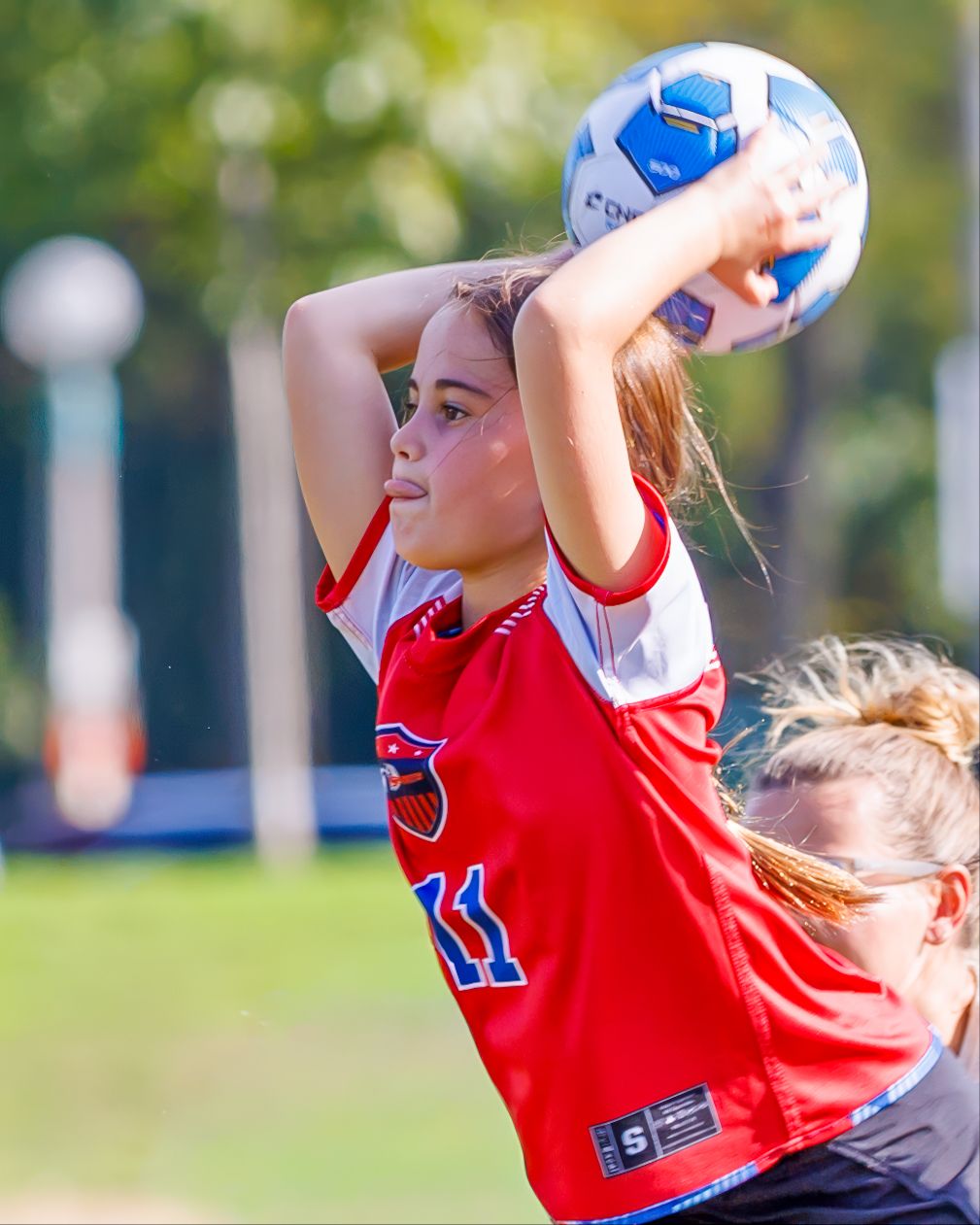 Female soccer player in red jersey #11 throws ball in during outdoor match on sunny day.