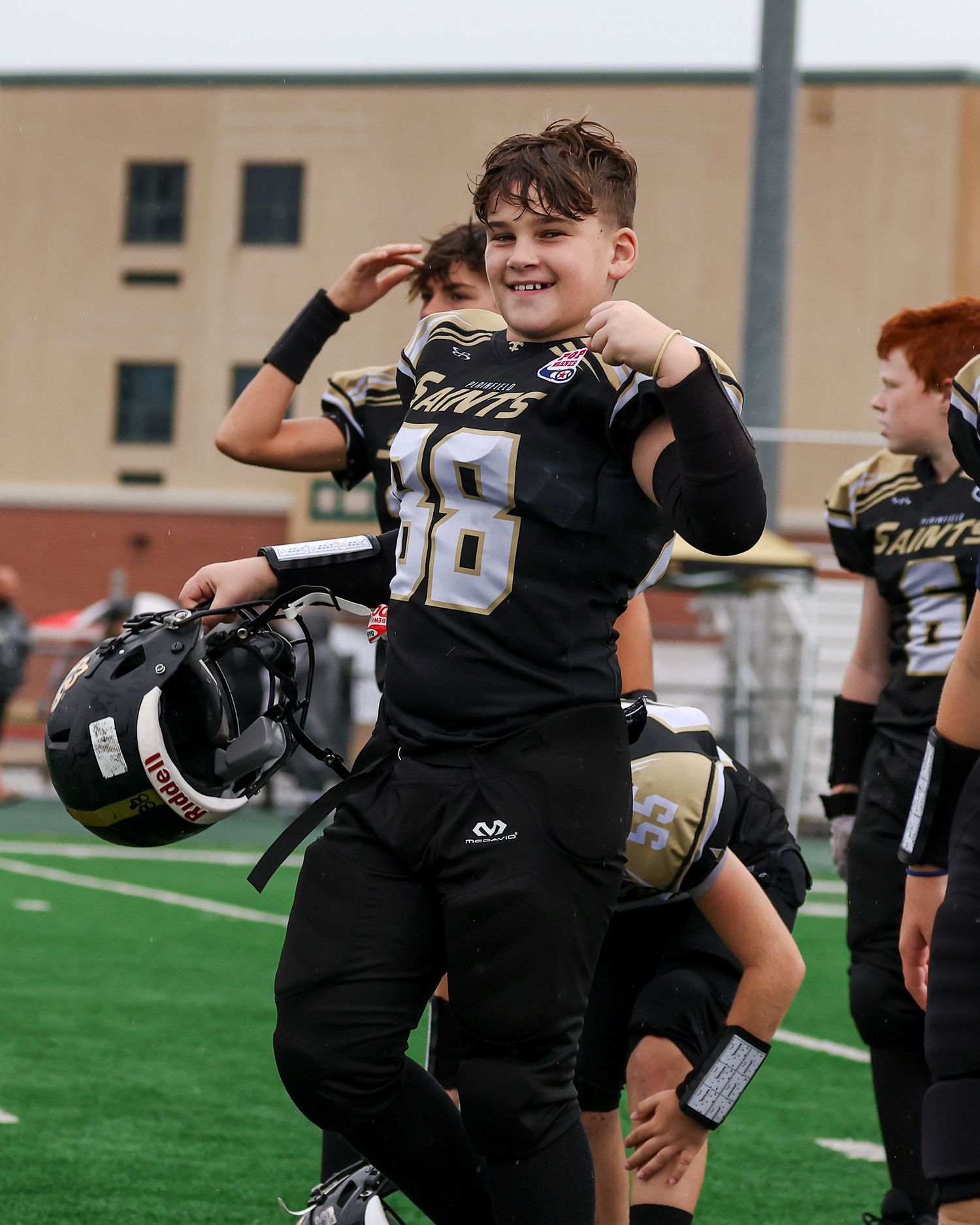 Young Saints football player smiling in black uniform, holding helmet on a green field during a youth football game.