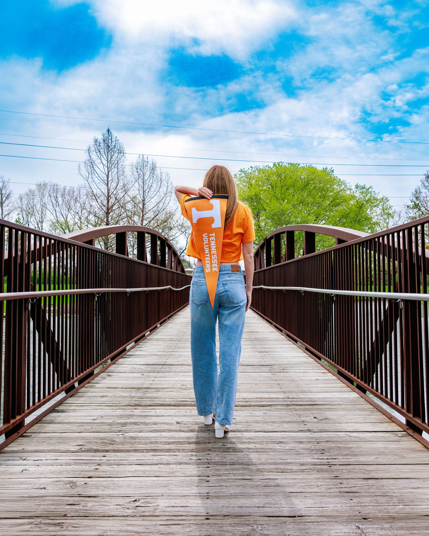 A young woman in an orange Tennessee shirt and wide-leg jeans walks across a wooden bridge under a blue sky.