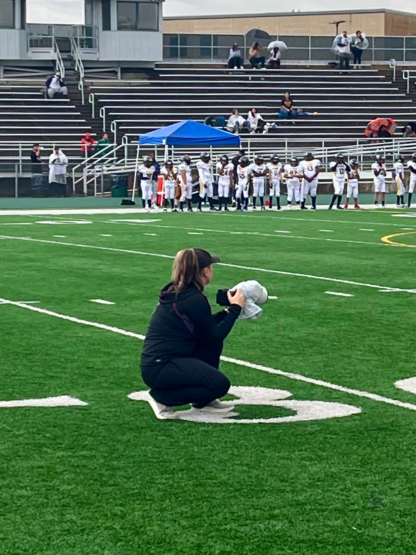 Sports photographer laying on artificial turf field capturing action shots during a football game with bleachers visible.