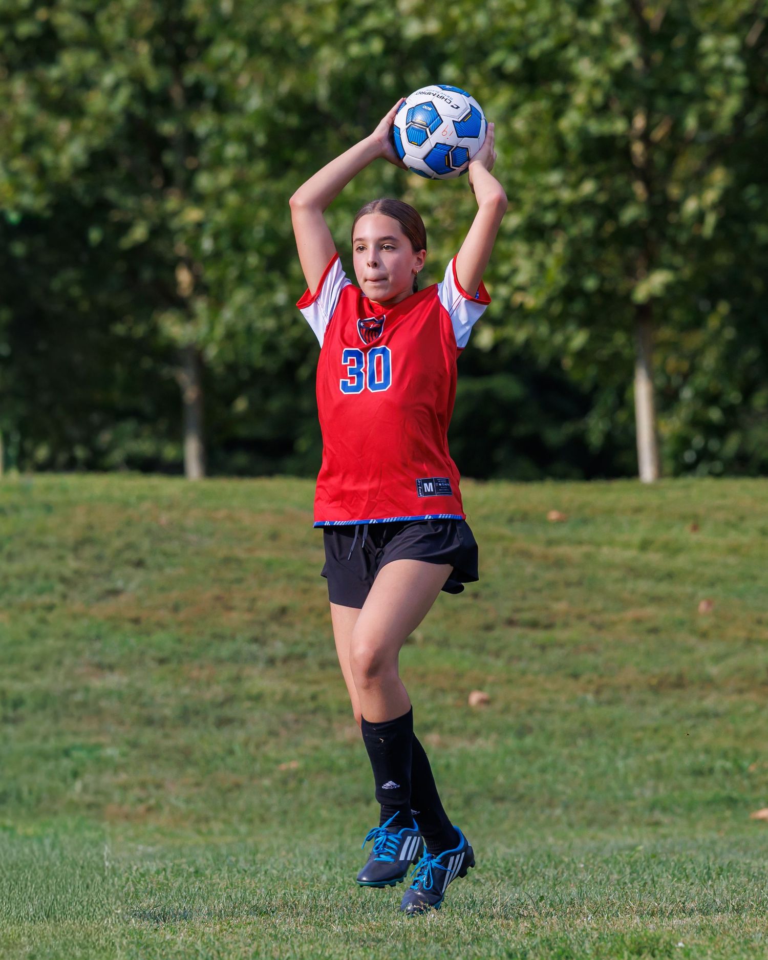 Girl in red jersey #30 performs a soccer throw-in on a green field during a youth soccer game.