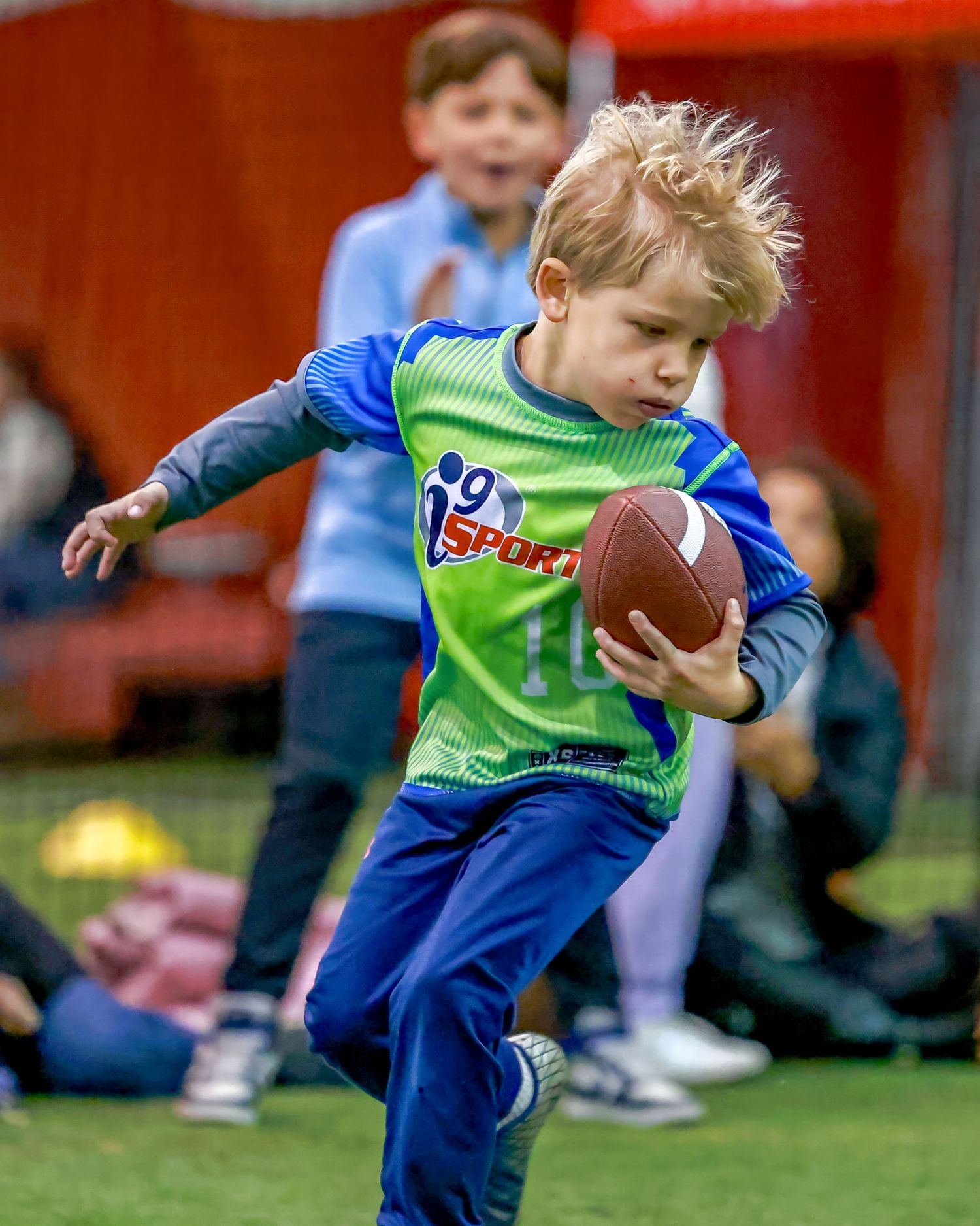 Young flag football player in green jersey runs with football on indoor field.