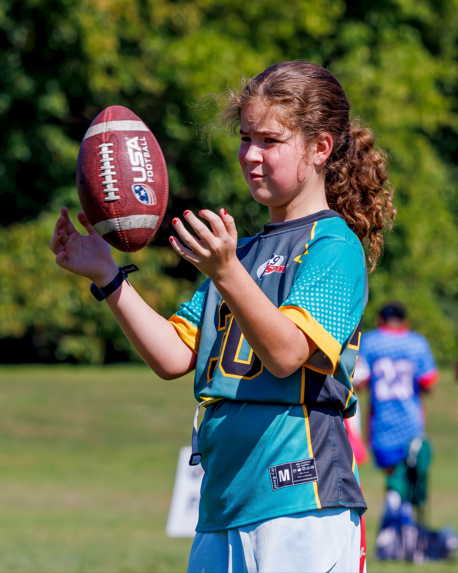 A player in a teal jersey practices catching a football outdoors on a sunny day.