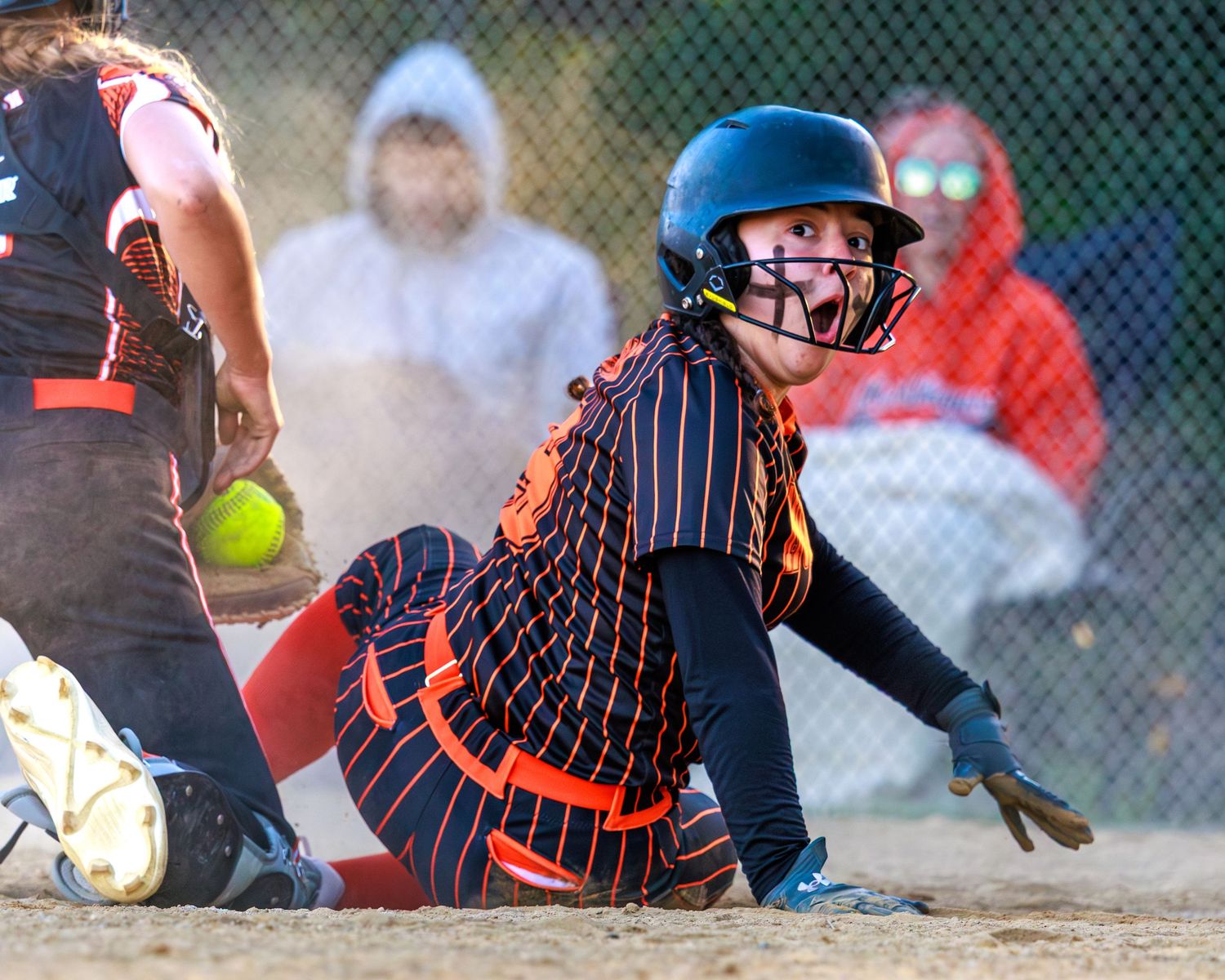 Softball player in orange uniform slides into base as ball approaches during an intense game play.