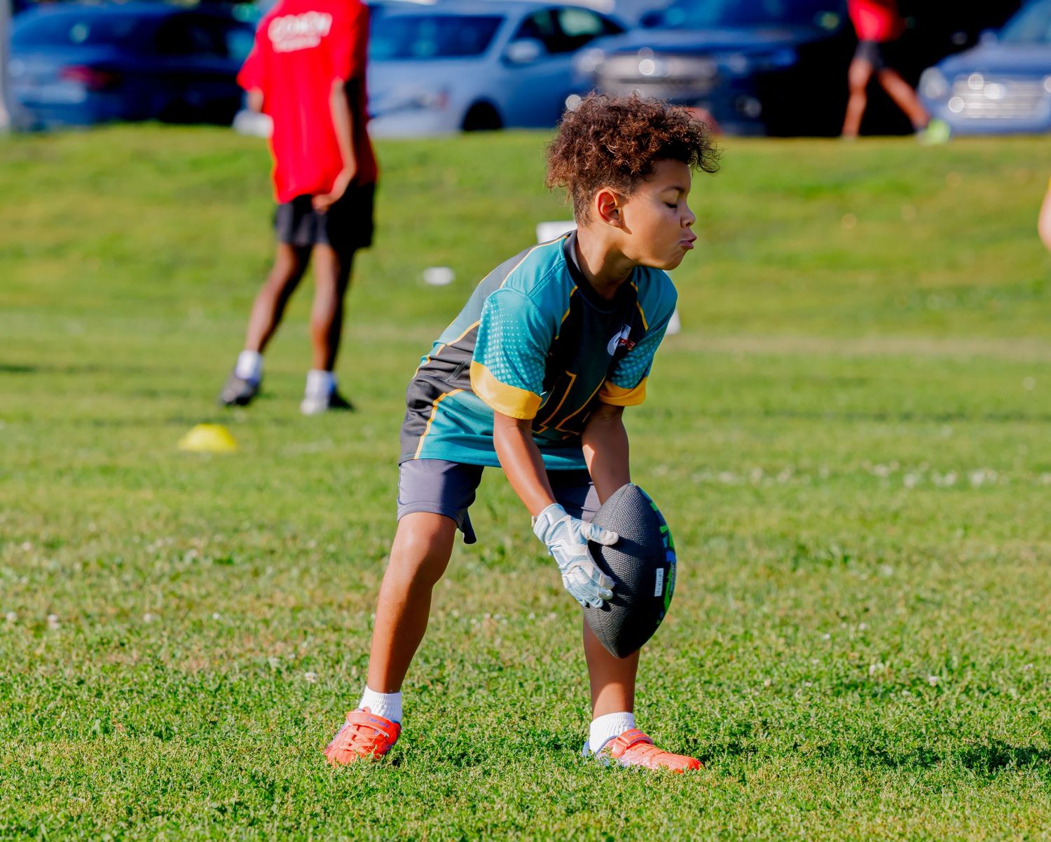 Young boy in teal jersey catches football during youth flag football practice on green grass field.