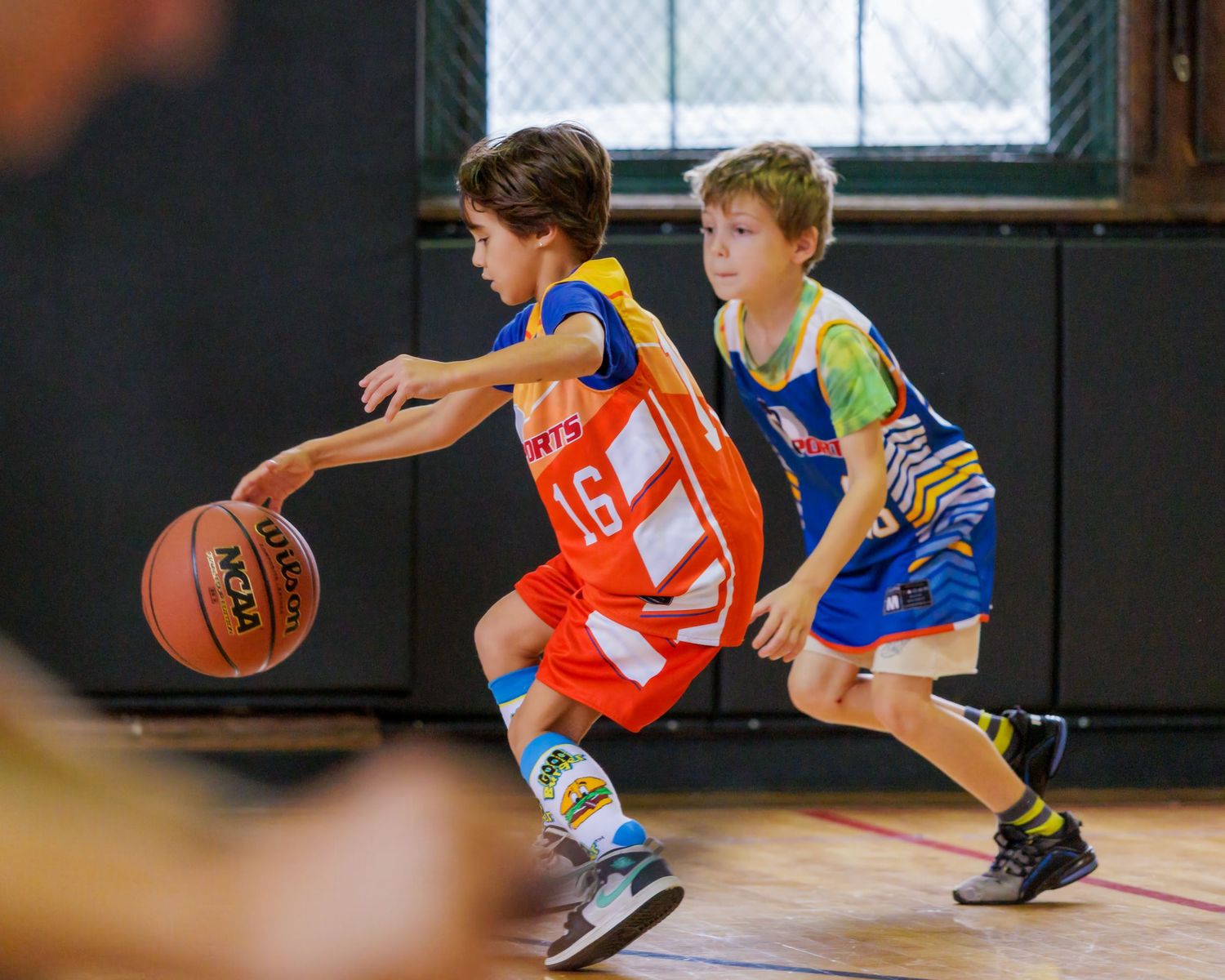 Two young boys playing basketball in a gym, one dribbling in an orange jersey numbered 16.