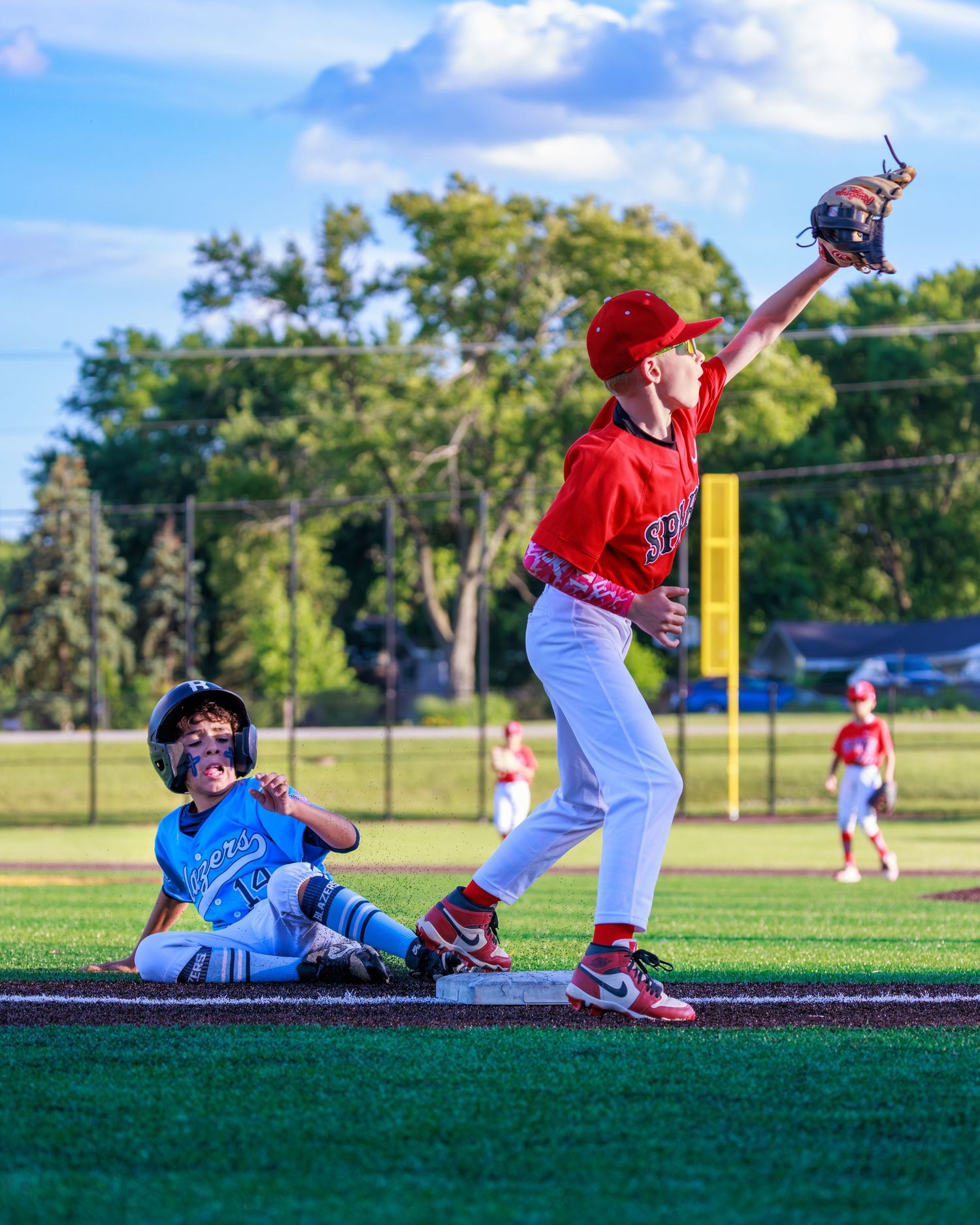 Youth baseball players in action, red-uniformed fielder catches ball as blue-uniformed runner slides into base.