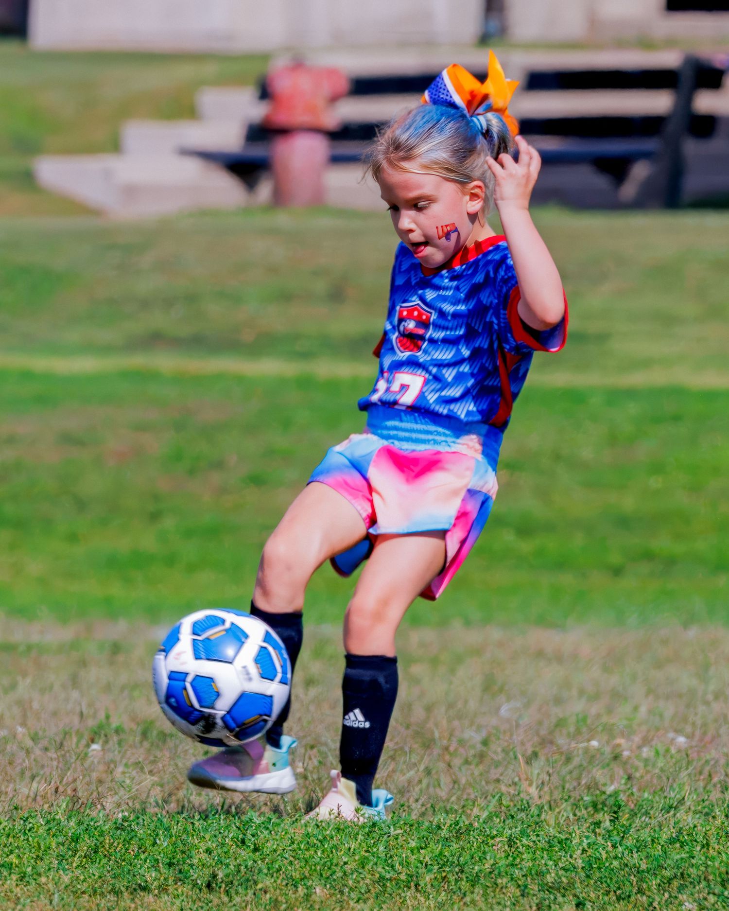 Young girl in blue soccer jersey kicking a ball on green grass field during youth soccer game.
