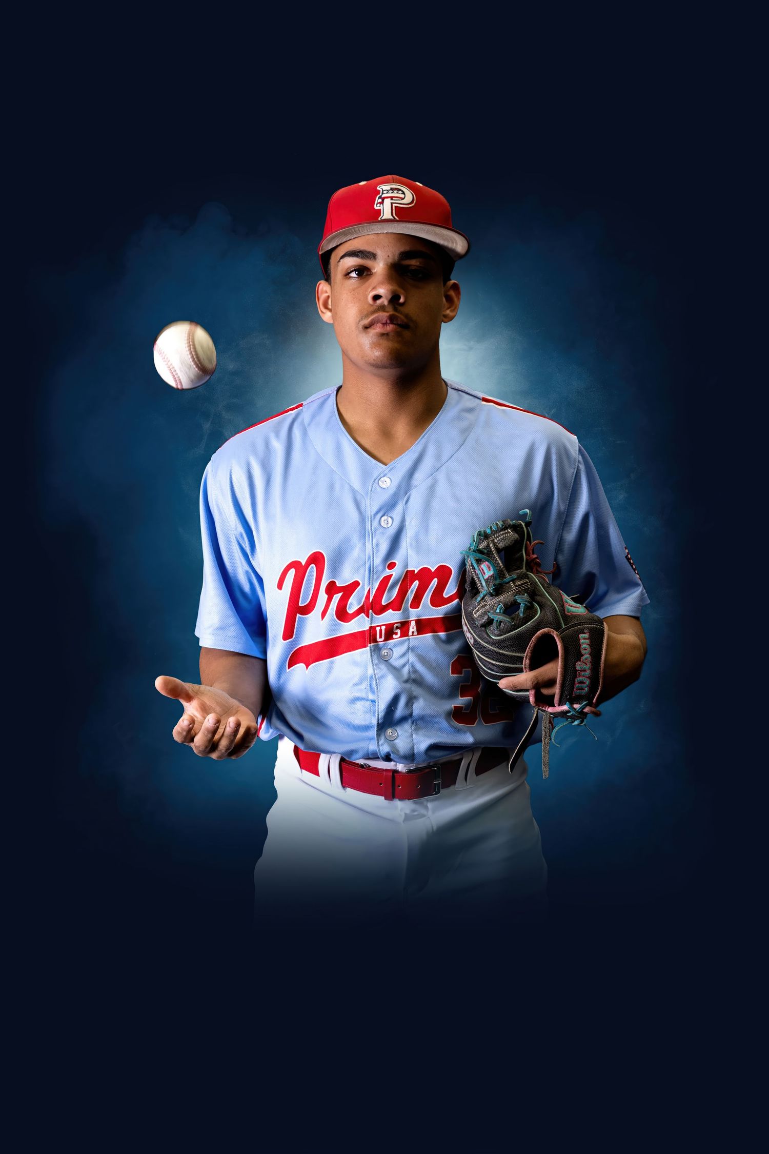 Young baseball player in Prism uniform tossing a ball while wearing a mitt, posed against a dark blue background.