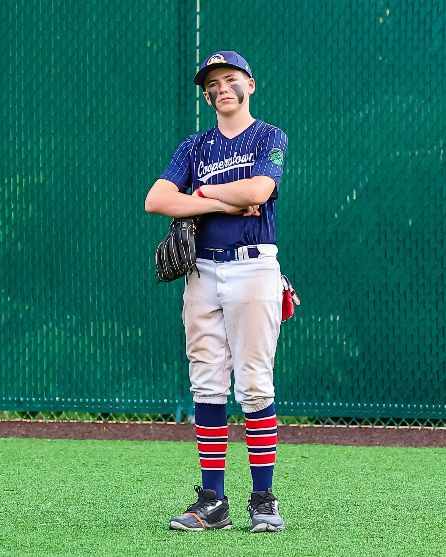 A young baseball player in white uniform with red and blue striped socks stands on a field against a green backdrop.