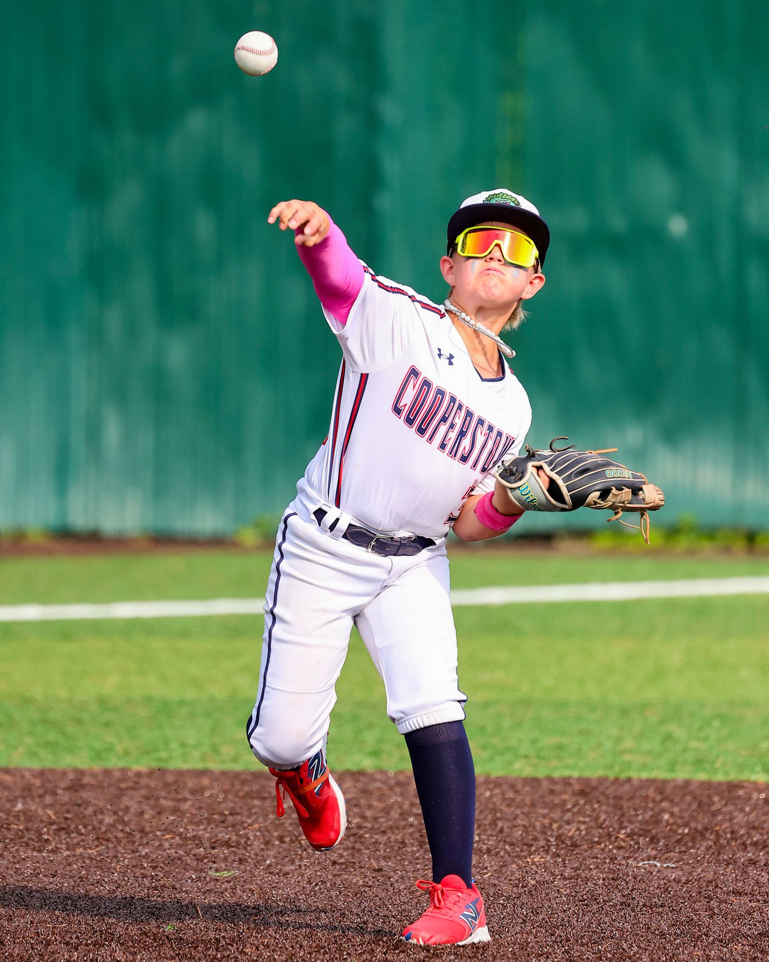 A baseball player in a white uniform with red accents reaches to catch a flying baseball against a green background.