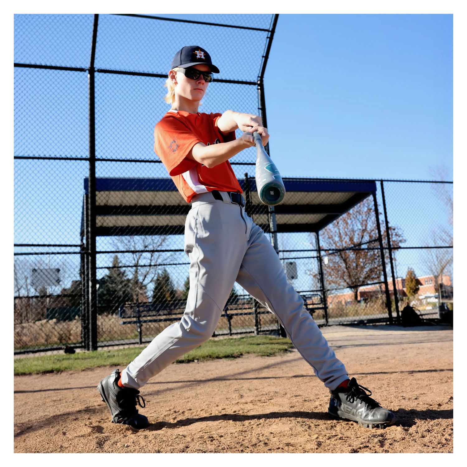 Baseball player in orange jersey demonstrates batting stance on field.