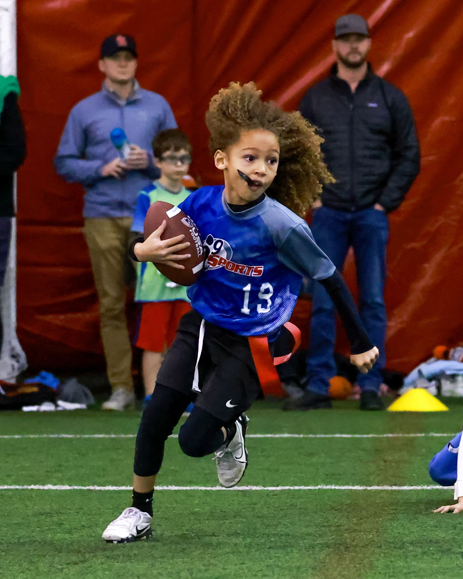 Youth flag football player in blue jersey number 13 runs with the ball during indoor game.