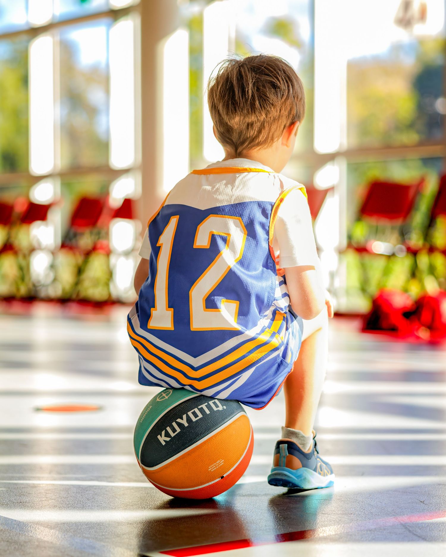 Young boy in blue number 12 basketball jersey crouching over a Kuyoto basketball on an indoor court.