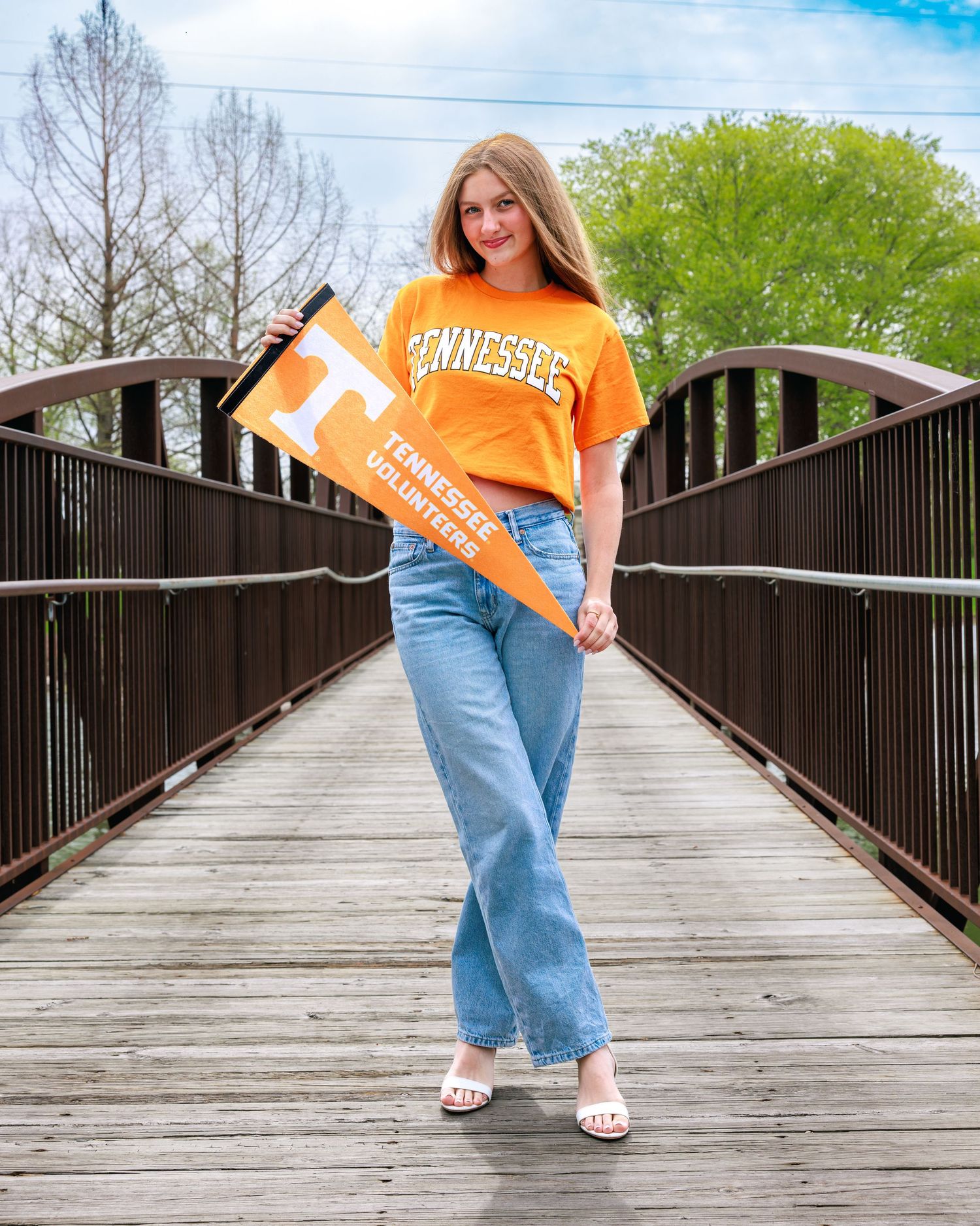Young woman in Tennessee Volunteers orange shirt and jeans holding a pennant on a wooden bridge.