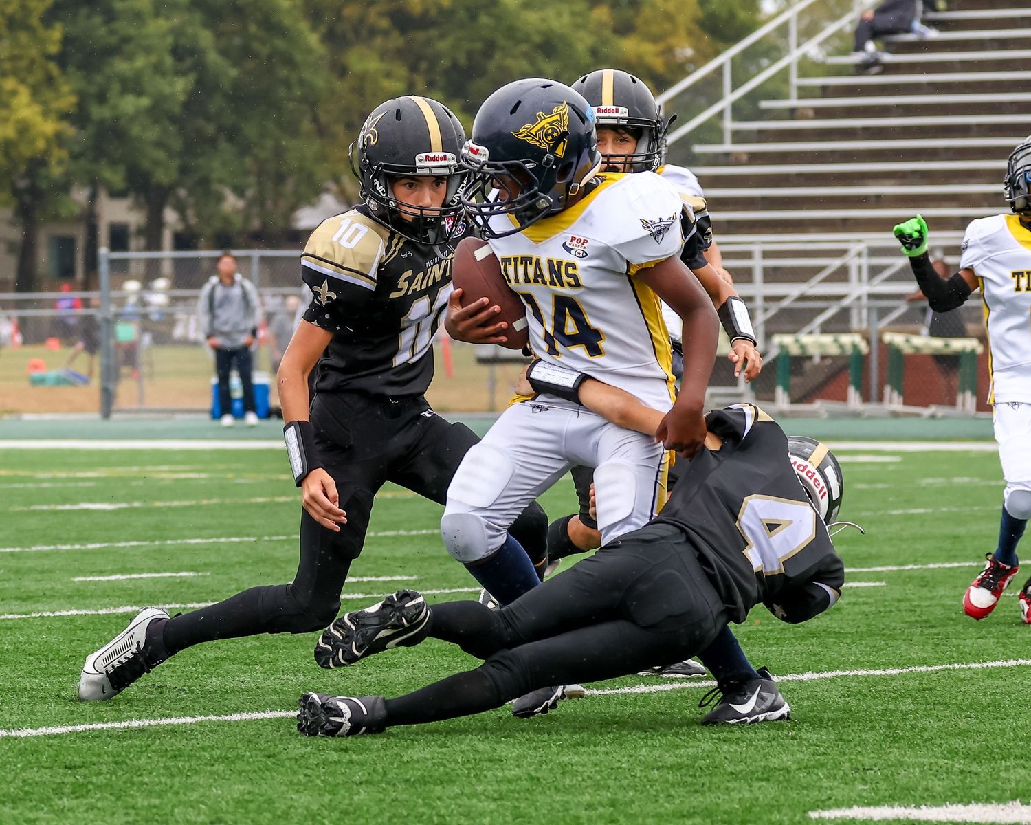 Youth football players in black and gold uniforms compete during a tackle play on a green turf field.