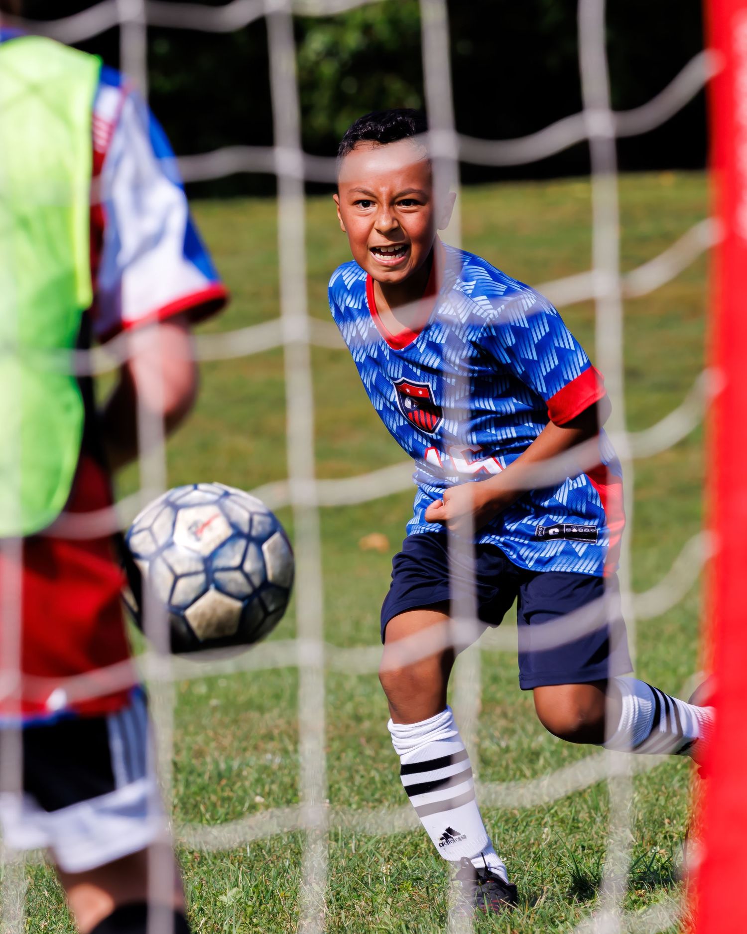 Soccer player in blue jersey and white socks crouches by goal post during match on sunny grass field.