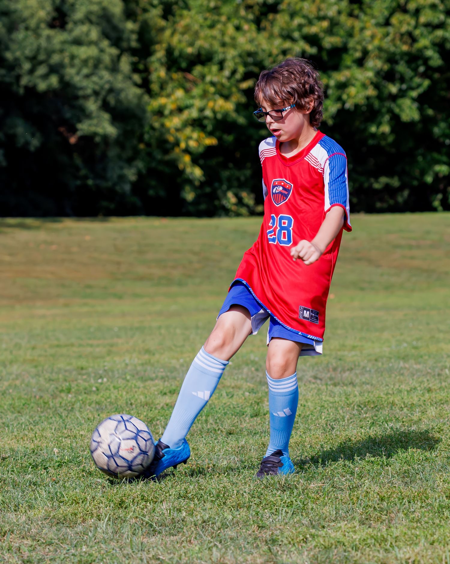 Young boy wearing glasses in red number 28 jersey kicking a soccer ball during a youth league game.