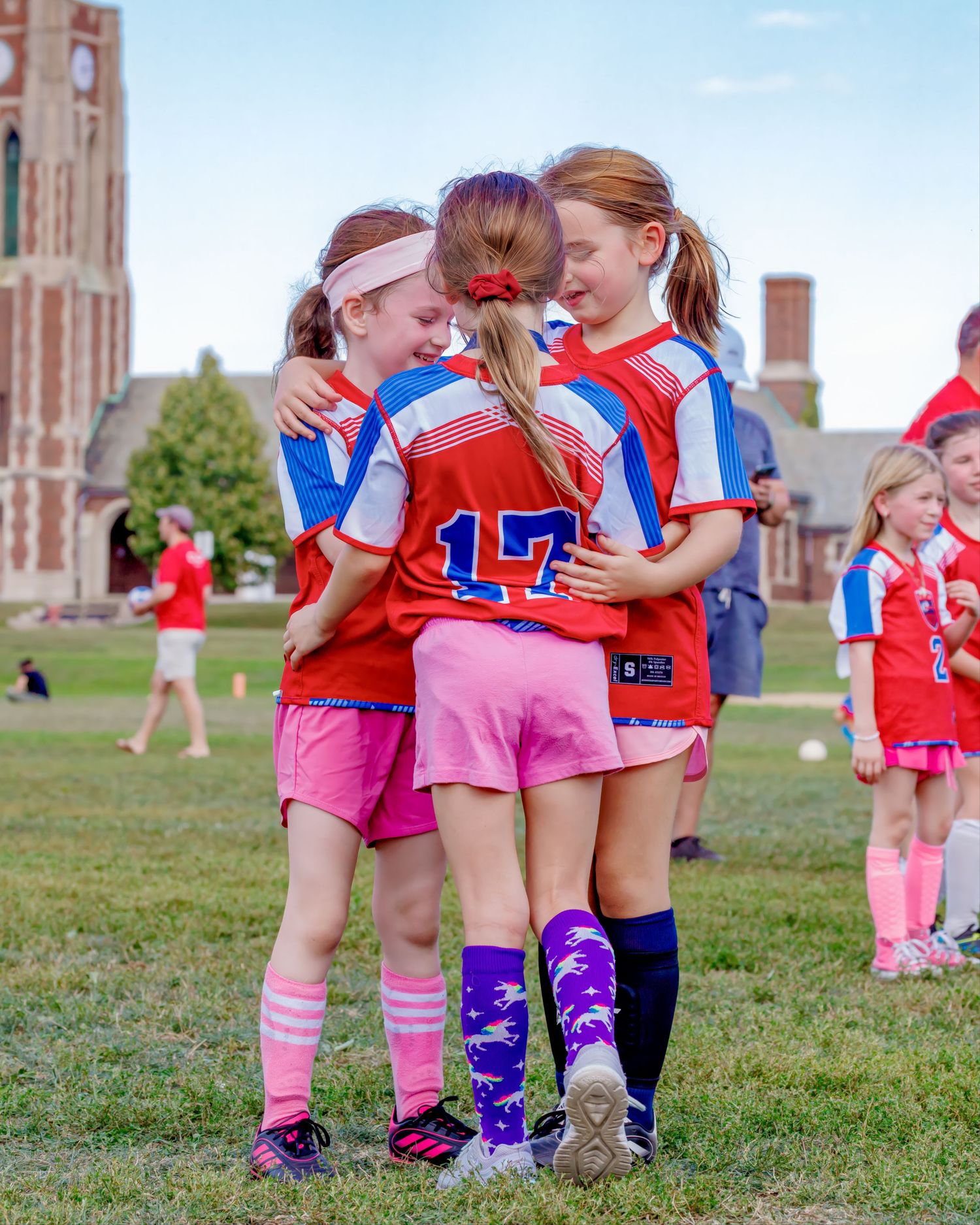 Three young girls in red soccer uniforms hugging on a grass field during a youth soccer game.