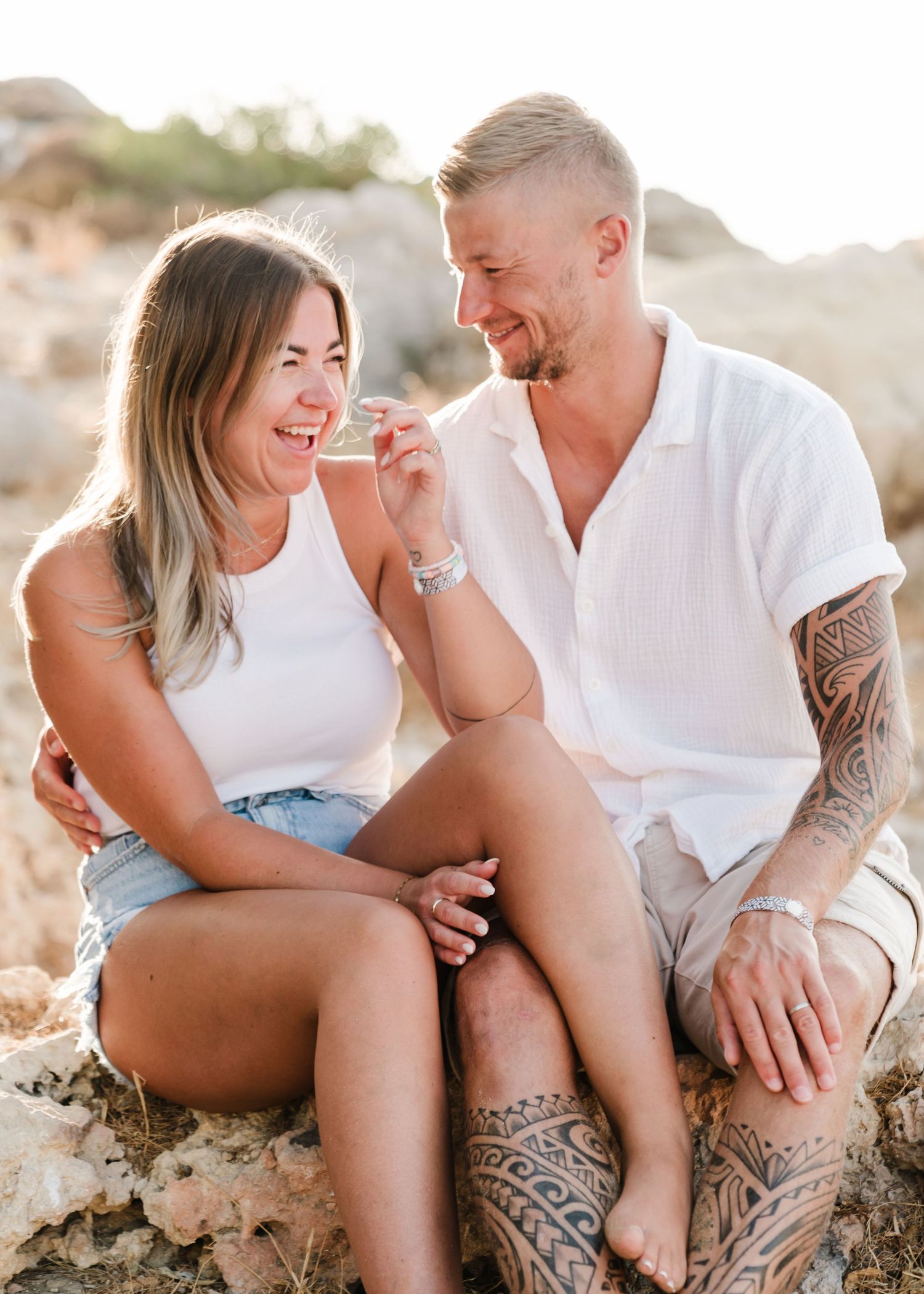 Joyful couple laughing together while sitting on rocky terrain in matching white tops. Fotograf Rhodos