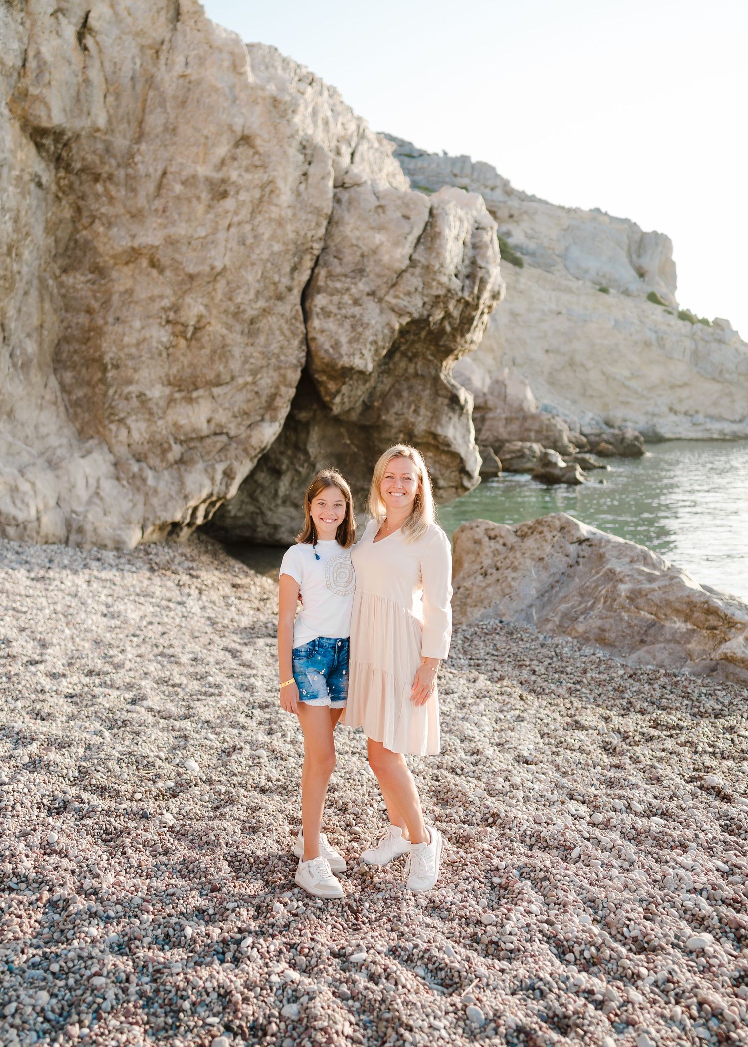 Two people stand together on a pebble beach with dramatic coastal cliffs and turquoise waters in the background. Fotograf Rhodos