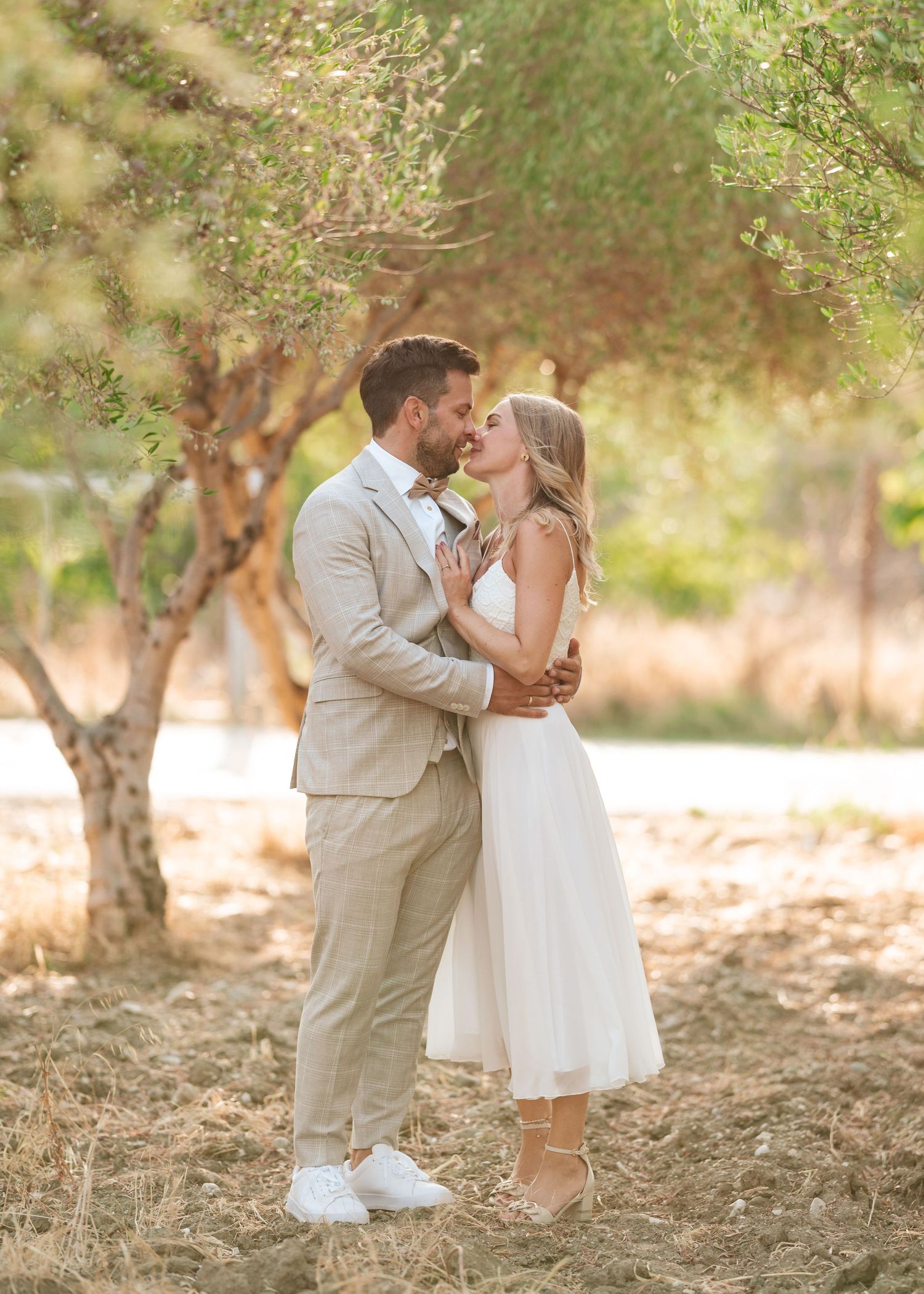 Ein Paar erlebt an seinem Hochzeitstag einen romantischen Moment in einem Olivenhain und trägt hellbeige und weiße Kleidung.